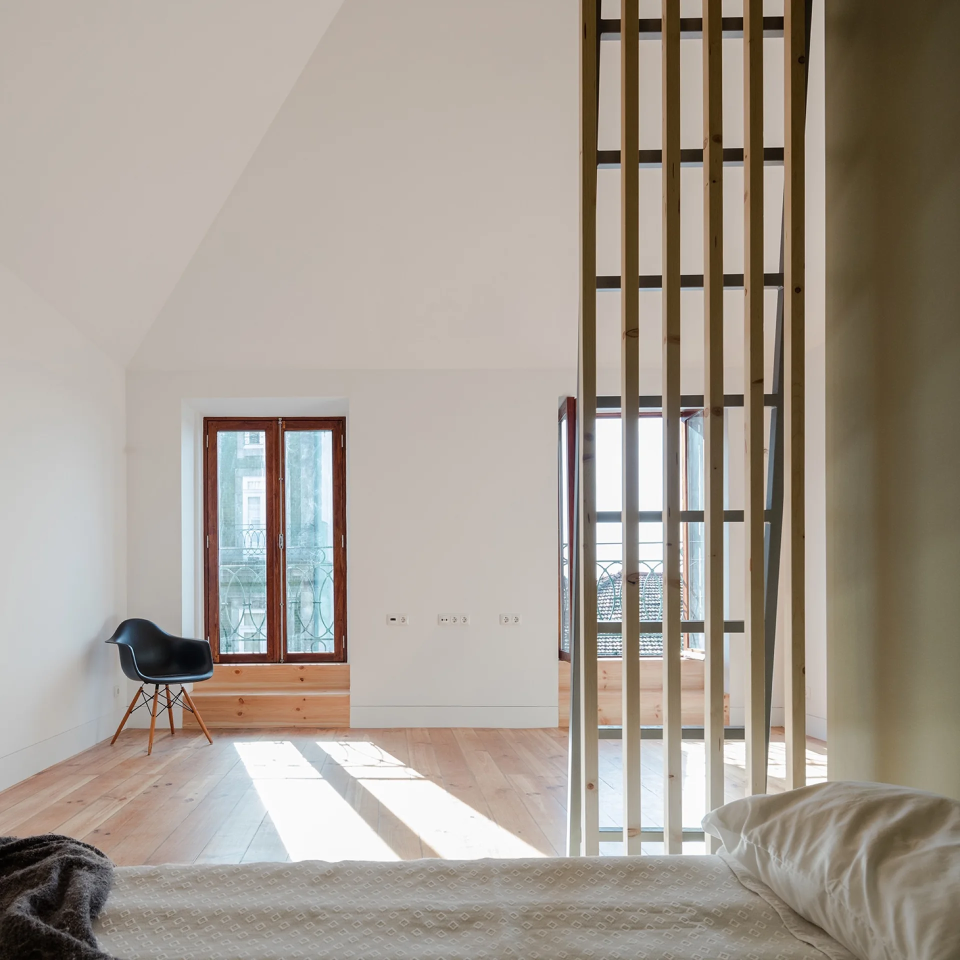 A minimalist bedroom features a wooden slat room divider next to a bed, overlooking a bright space with wood floors and a black chair.