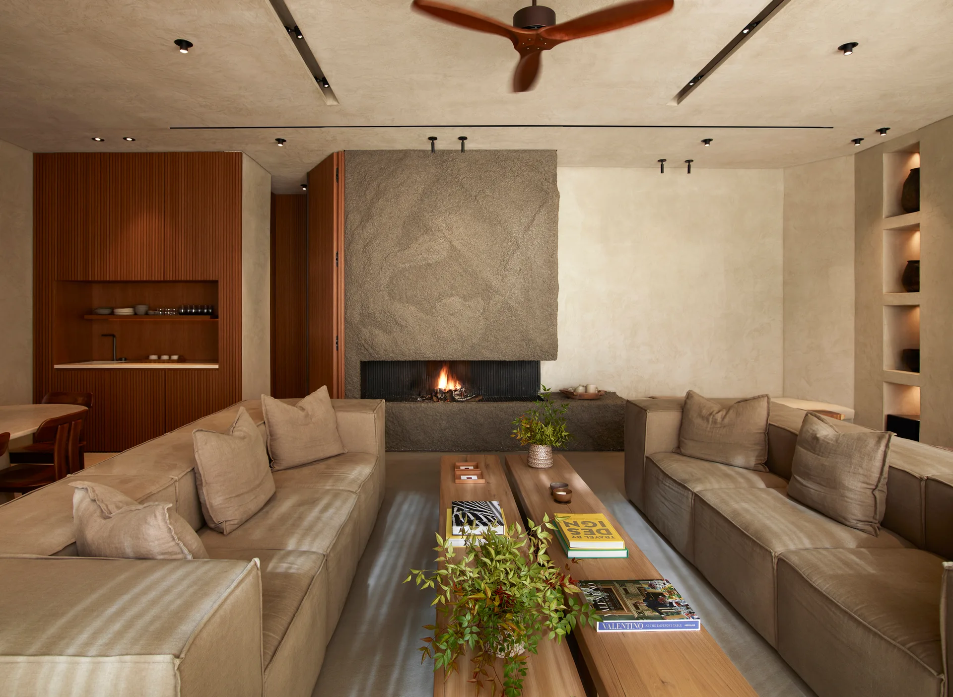 A minimalist living room features two neutral sofas facing long wood coffee tables in front of a massive textured stone fireplace.
