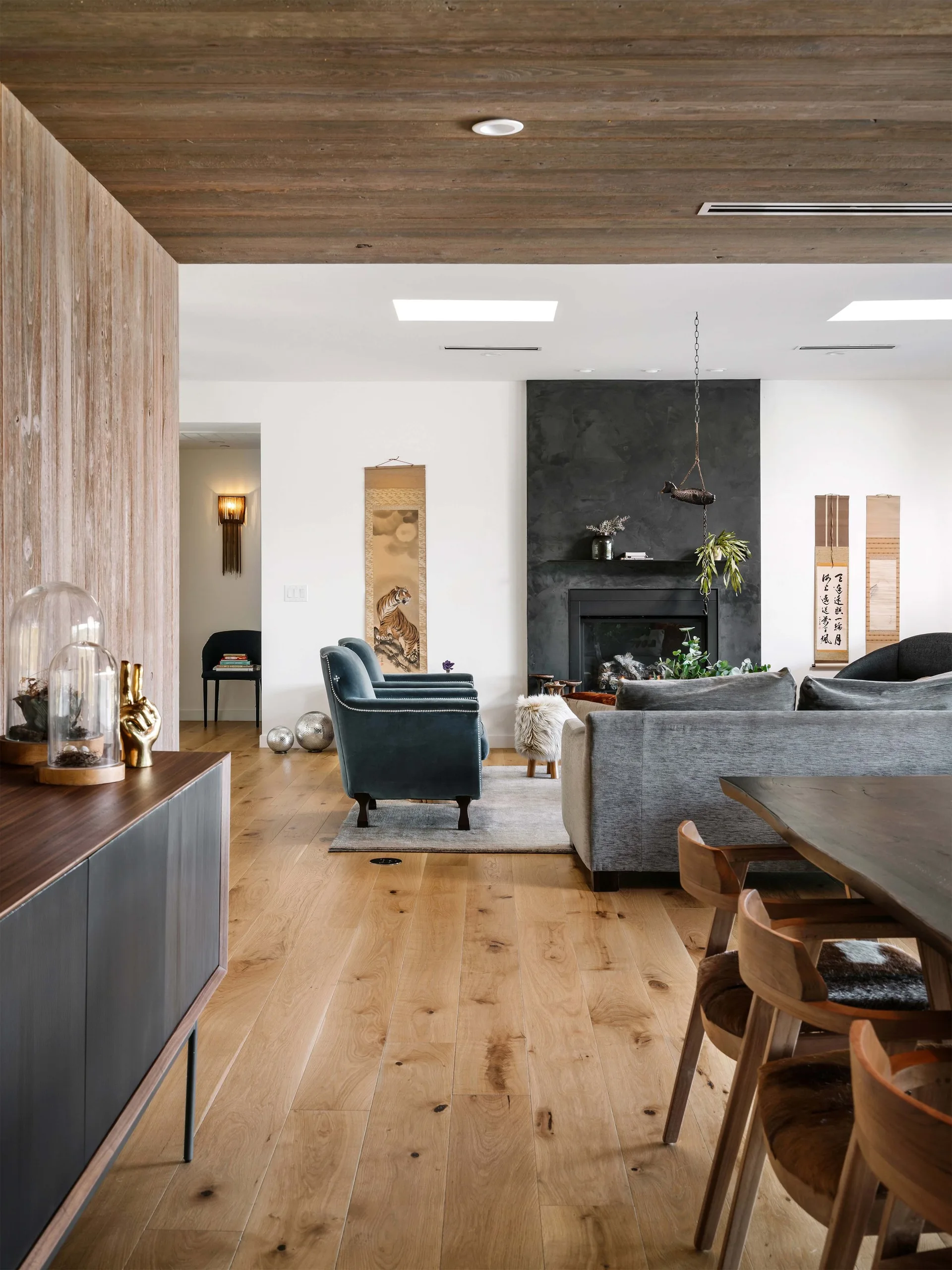 A modern living room with a wood plank ceiling, a matte black fireplace surround, a grey sofa, and teal velvet armchairs.