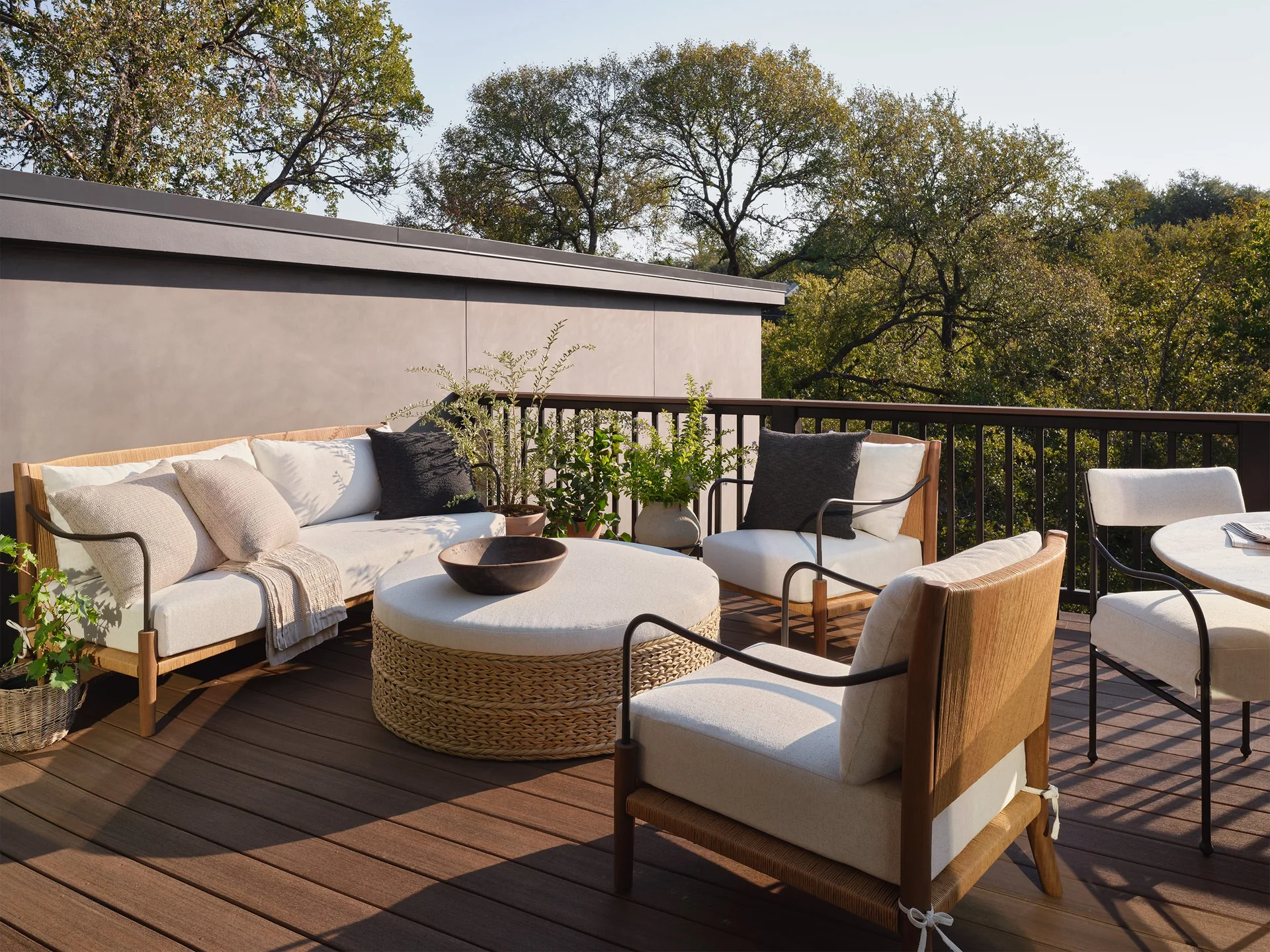 An outdoor wooden deck features a white cushioned sofa, two matching armchairs, and a round woven coffee table surrounded by green trees.