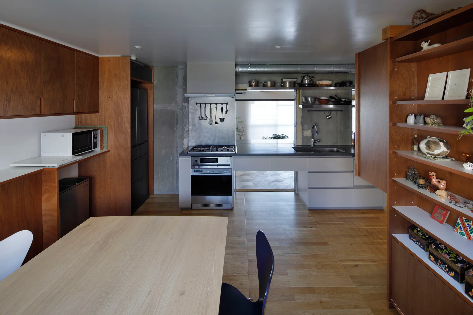 A modern kitchen features warm wood cabinetry, a raw concrete backsplash wall, stainless steel appliances, and a dining table in the foreground.
