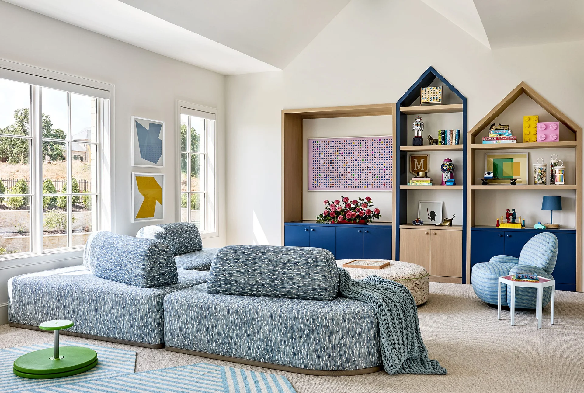 A bright kids' playroom featuring a patterned blue modular sofa, house-shaped built-in shelving with toys, and large windows.