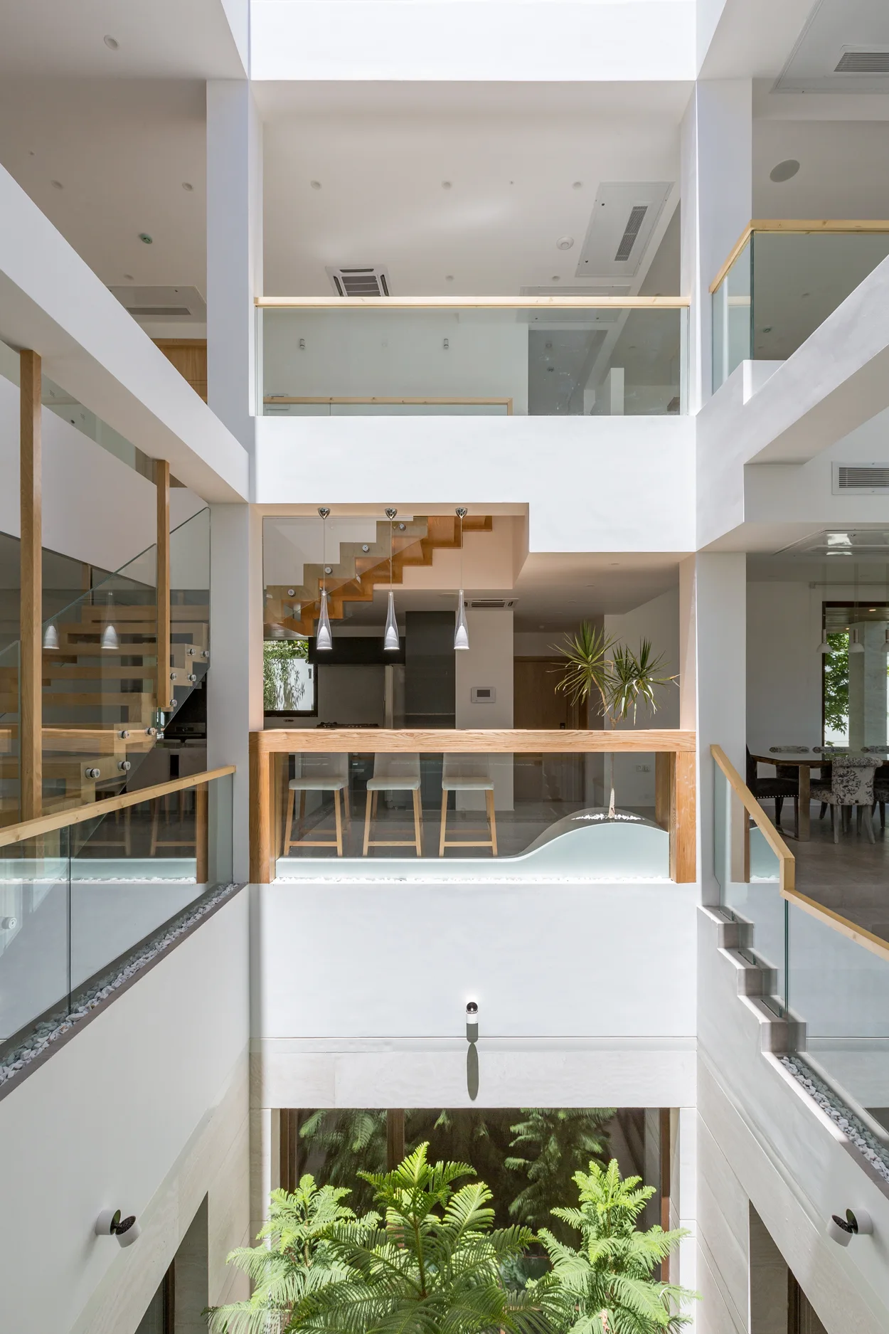 A white multi-level interior atrium featuring glass and wood railings, open wooden staircases, and green plants at the bottom.