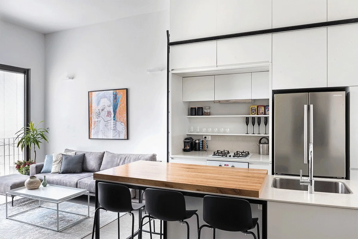 A minimalist open-concept kitchen features a white island with a thick wood countertop, black bar stools, and a grey sectional sofa in the background.