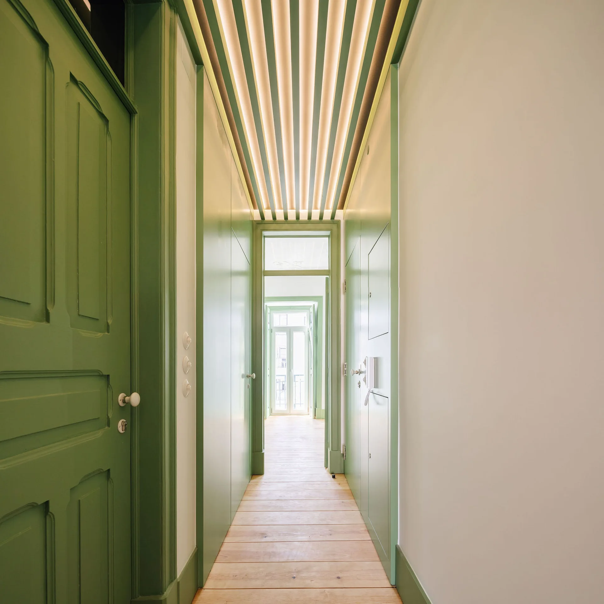 A long narrow hallway features sage green painted walls and doors, light wood floors, and a modern slatted ceiling with integrated warm lighting.