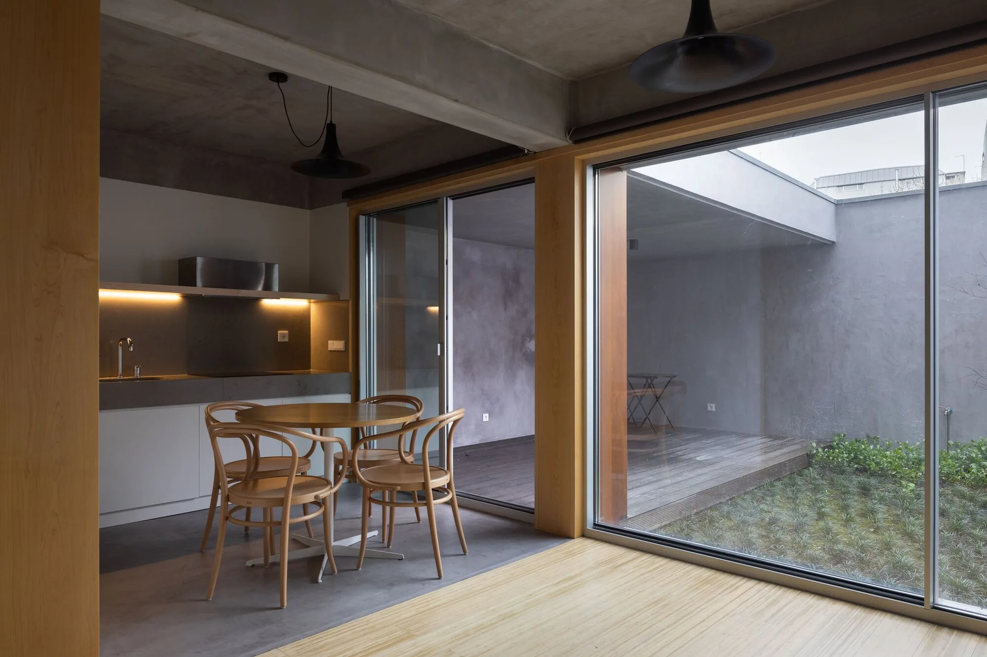 A minimalist dining area features a round wood table and bentwood chairs next to sliding glass doors that open to a concrete courtyard.