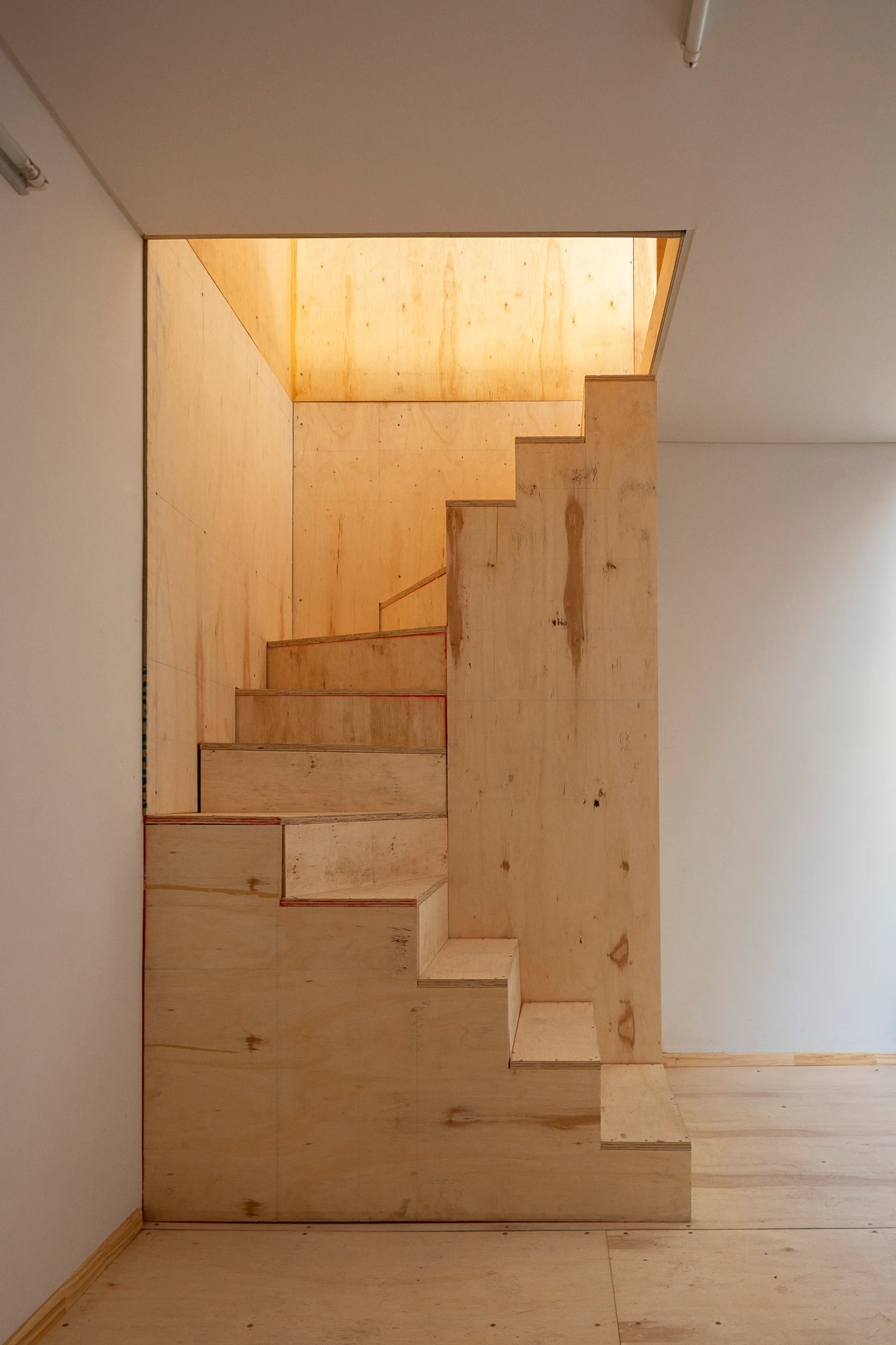 An unfinished plywood staircase rises into a bright opening within a minimalist white room with light wood flooring.
