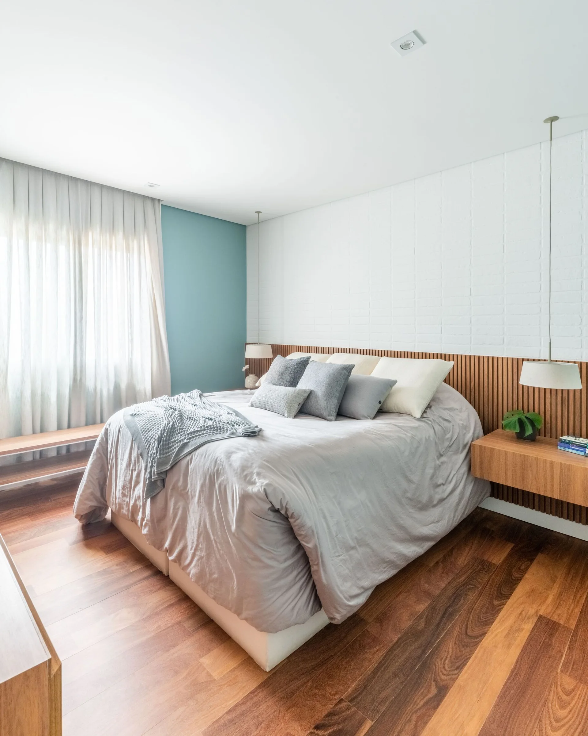A contemporary bedroom featuring a bed with grey linens against a slatted wood headboard panel and a white painted brick wall.