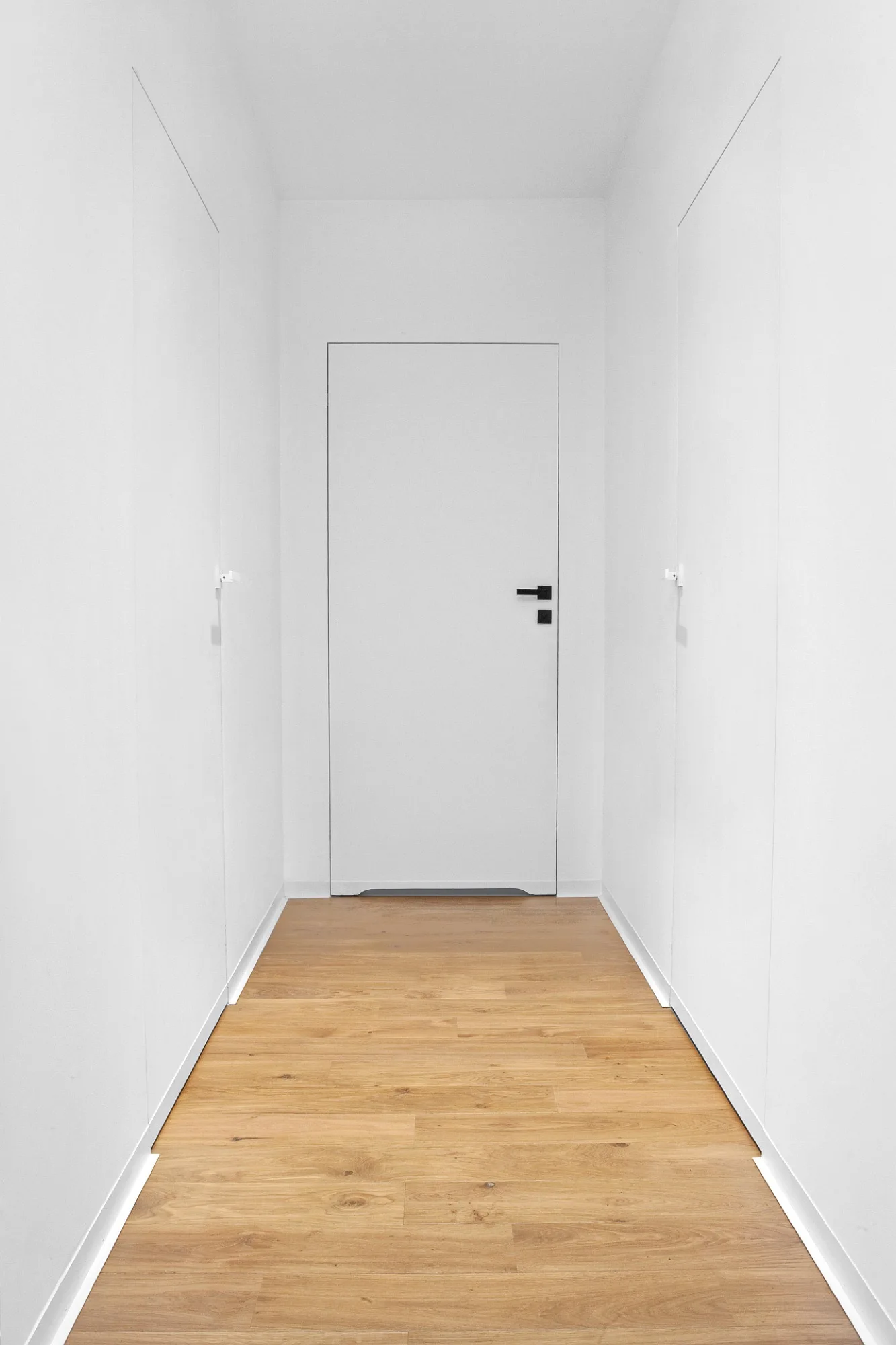 A symmetric white hallway features natural wood plank flooring and flush-to-wall doors with minimalist black hardware.