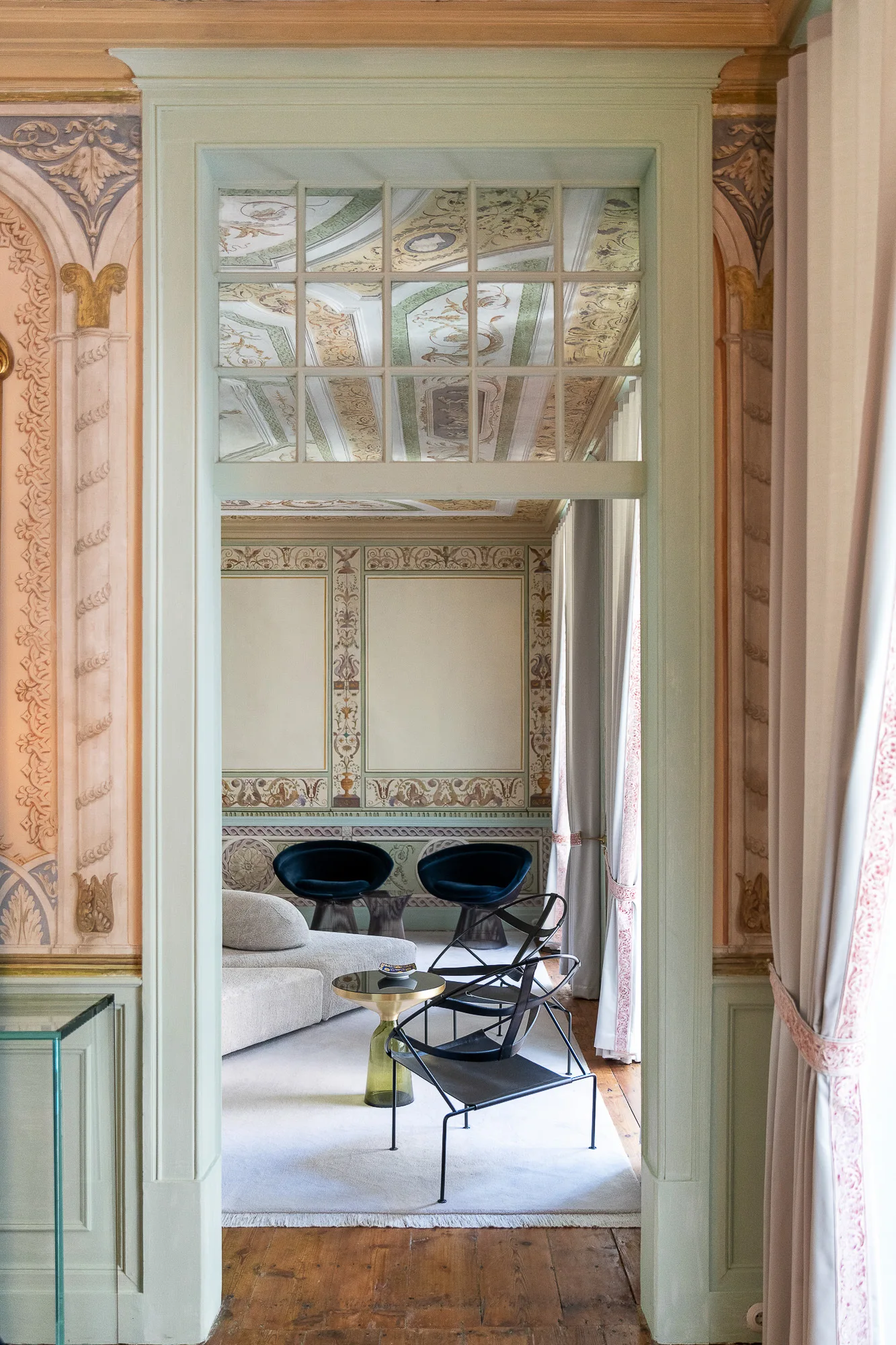 A view through a muted green doorway with a glass transom revealing a living room with ornate painted wall murals, a curved sofa, and modern chairs.
