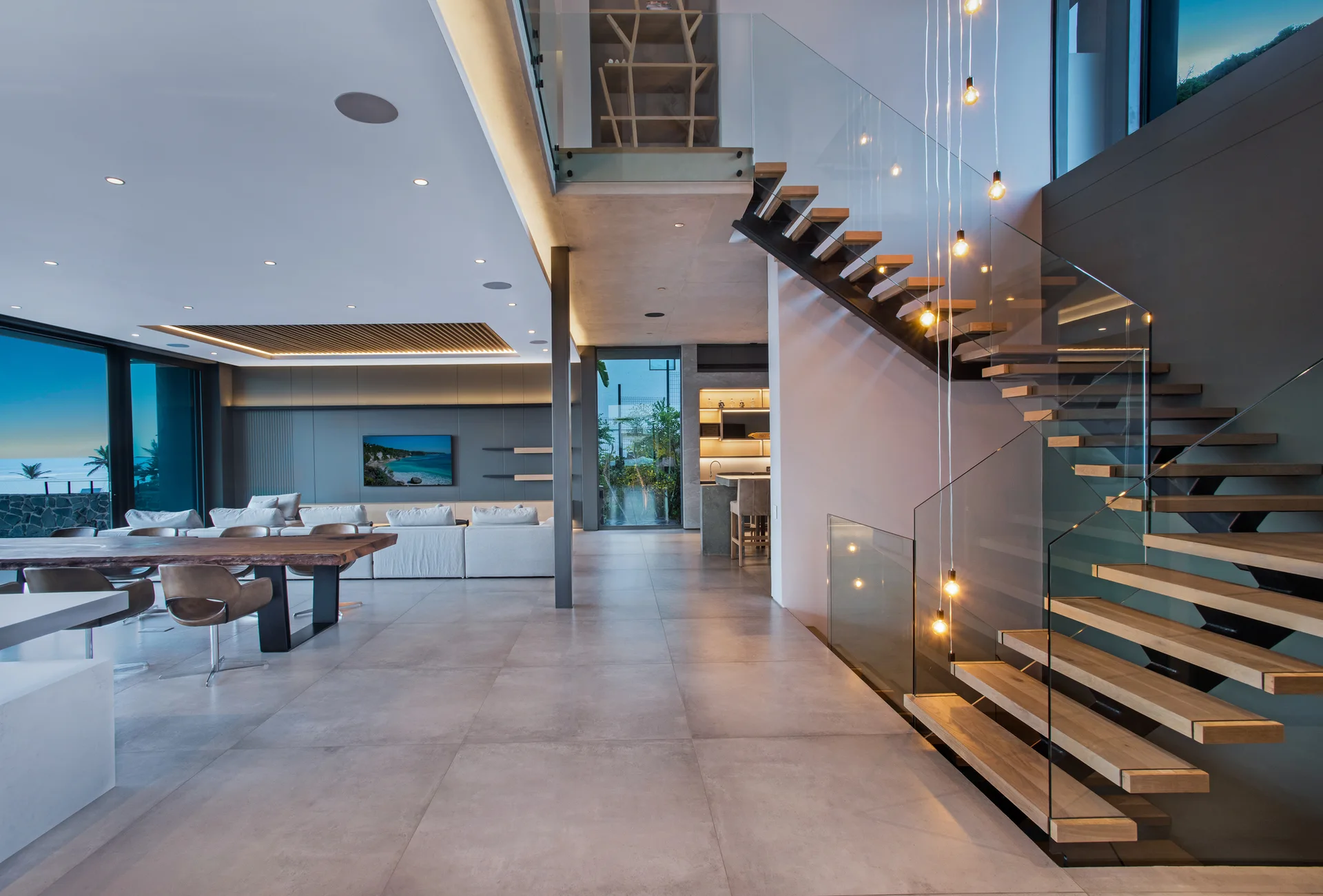 A modern open-plan living and dining space featuring a floating wooden staircase with a glass railing, a live-edge table, and large grey floor tiles.