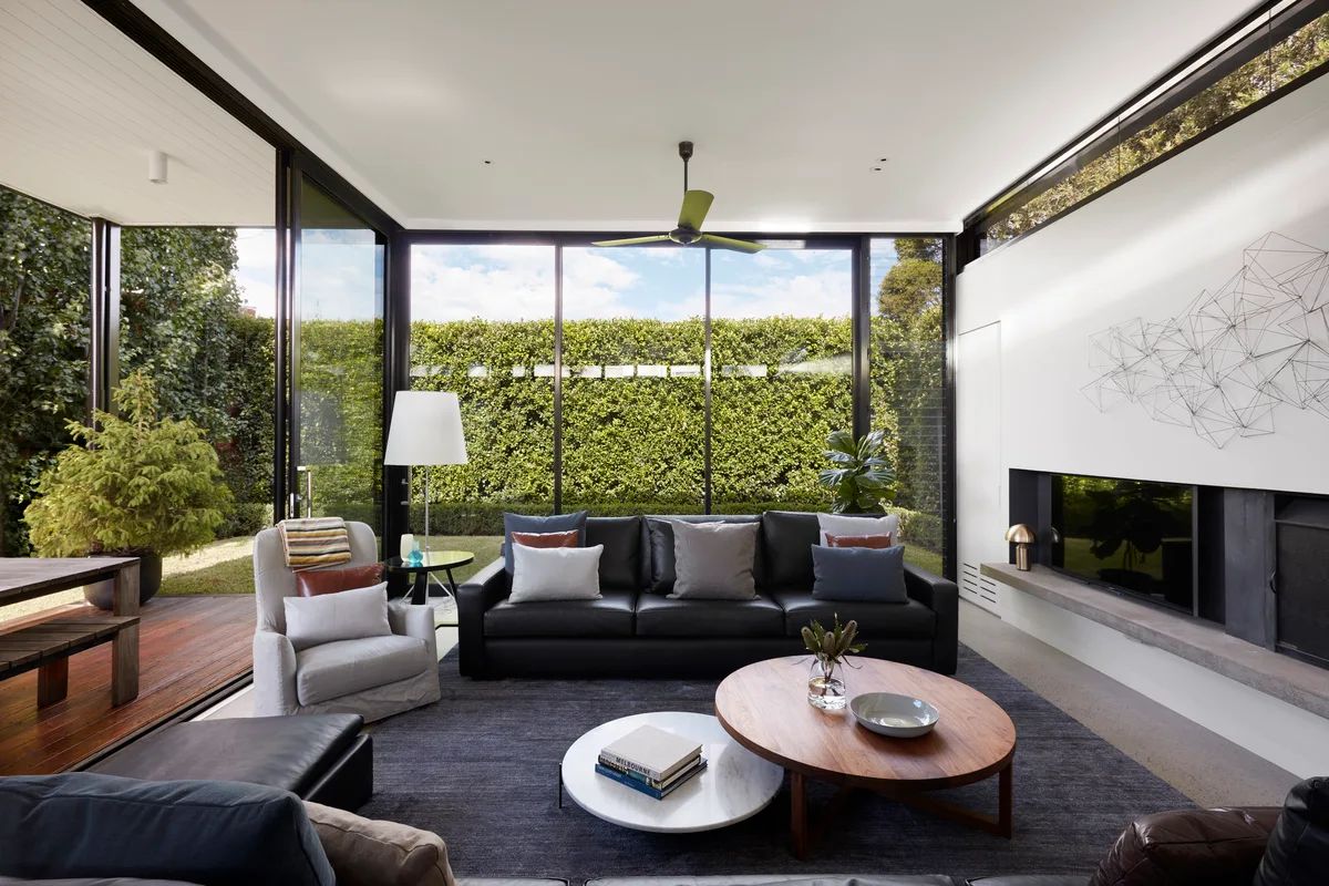 A modern living room with a black leather sofa, round wooden and marble coffee tables, and glass walls looking out to a tall hedge.