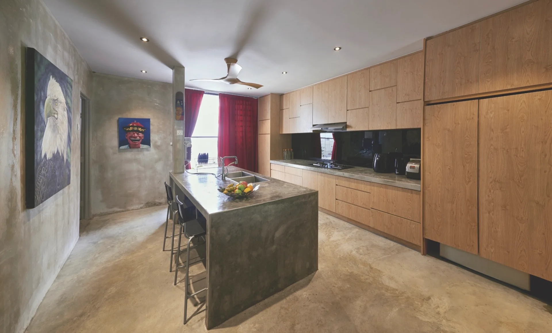 An industrial kitchen features a large concrete island with barstools, raw concrete walls, warm wood cabinetry, and a vibrant red curtain in the background.