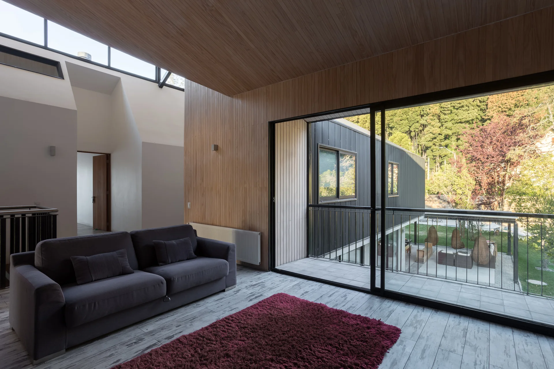 A modern living room with vertical wood paneling on the walls and ceiling, a grey sofa, and large sliding glass doors opening to a balcony.