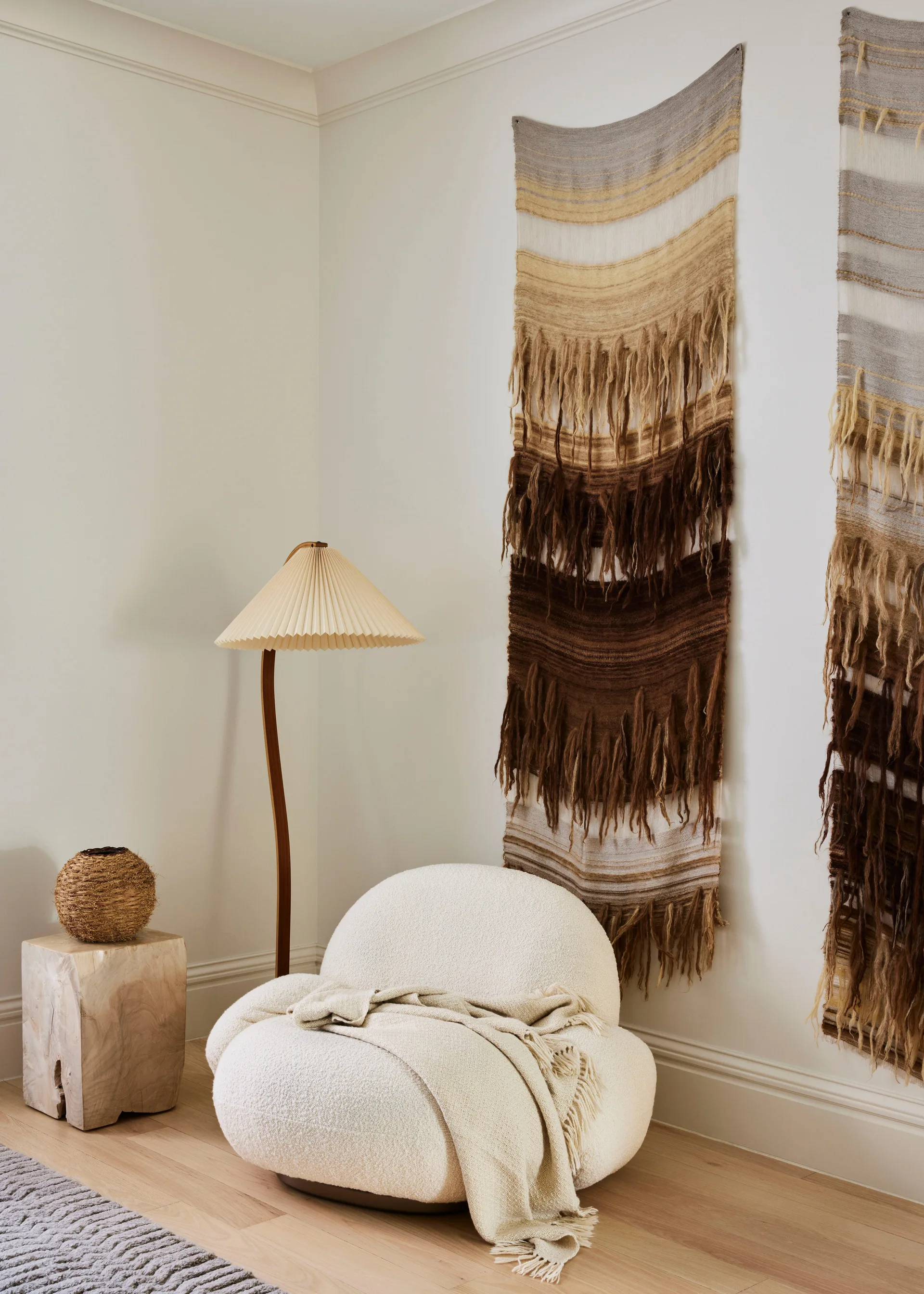 A cozy corner featuring a rounded white boucle armchair, a curved wood floor lamp with a pleated shade, and shaggy earth-toned wall tapestries.