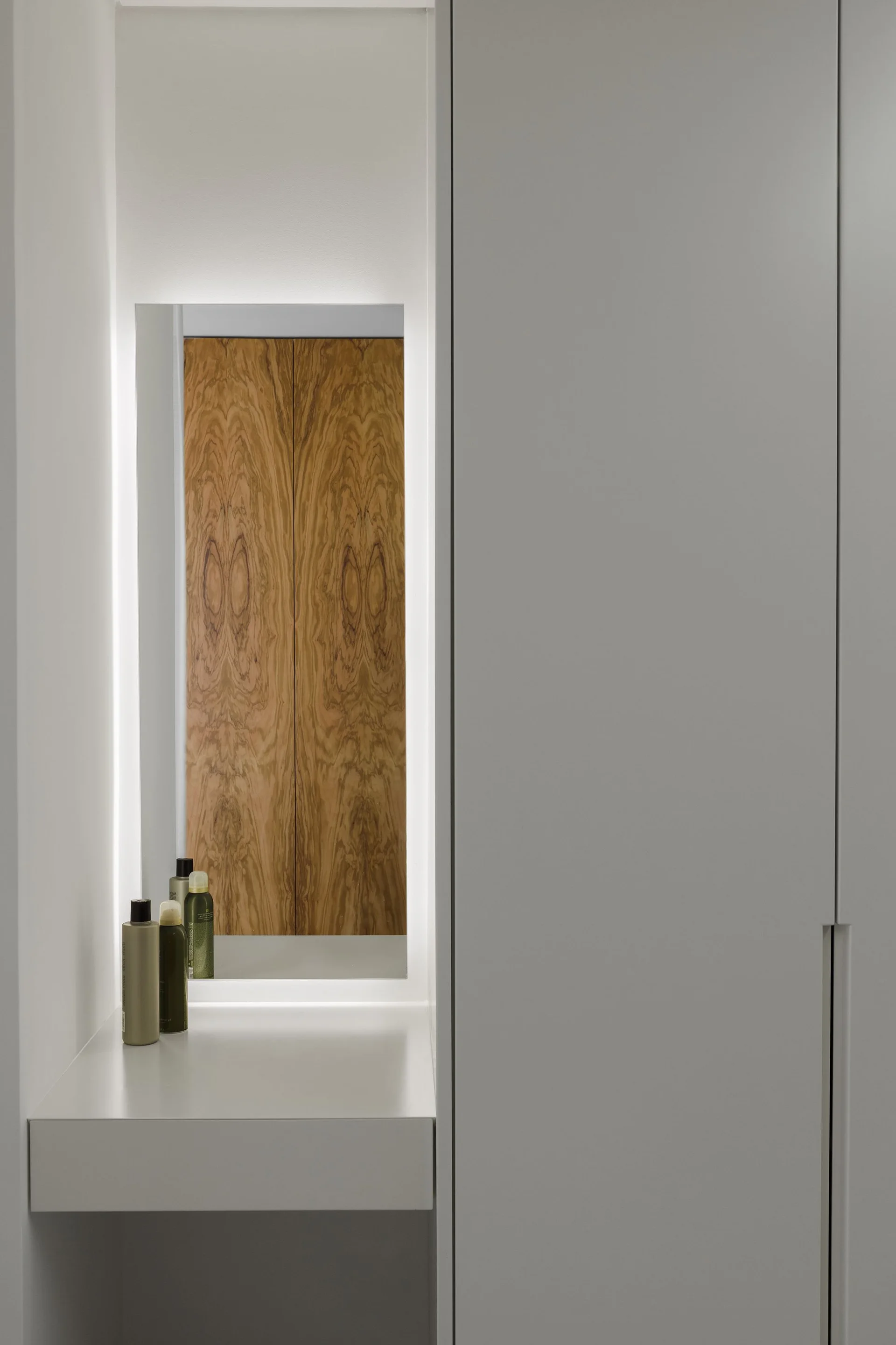 A minimalist white dressing area with a built-in vanity shelf, matte cabinets, and a backlit mirror reflecting wood grain.