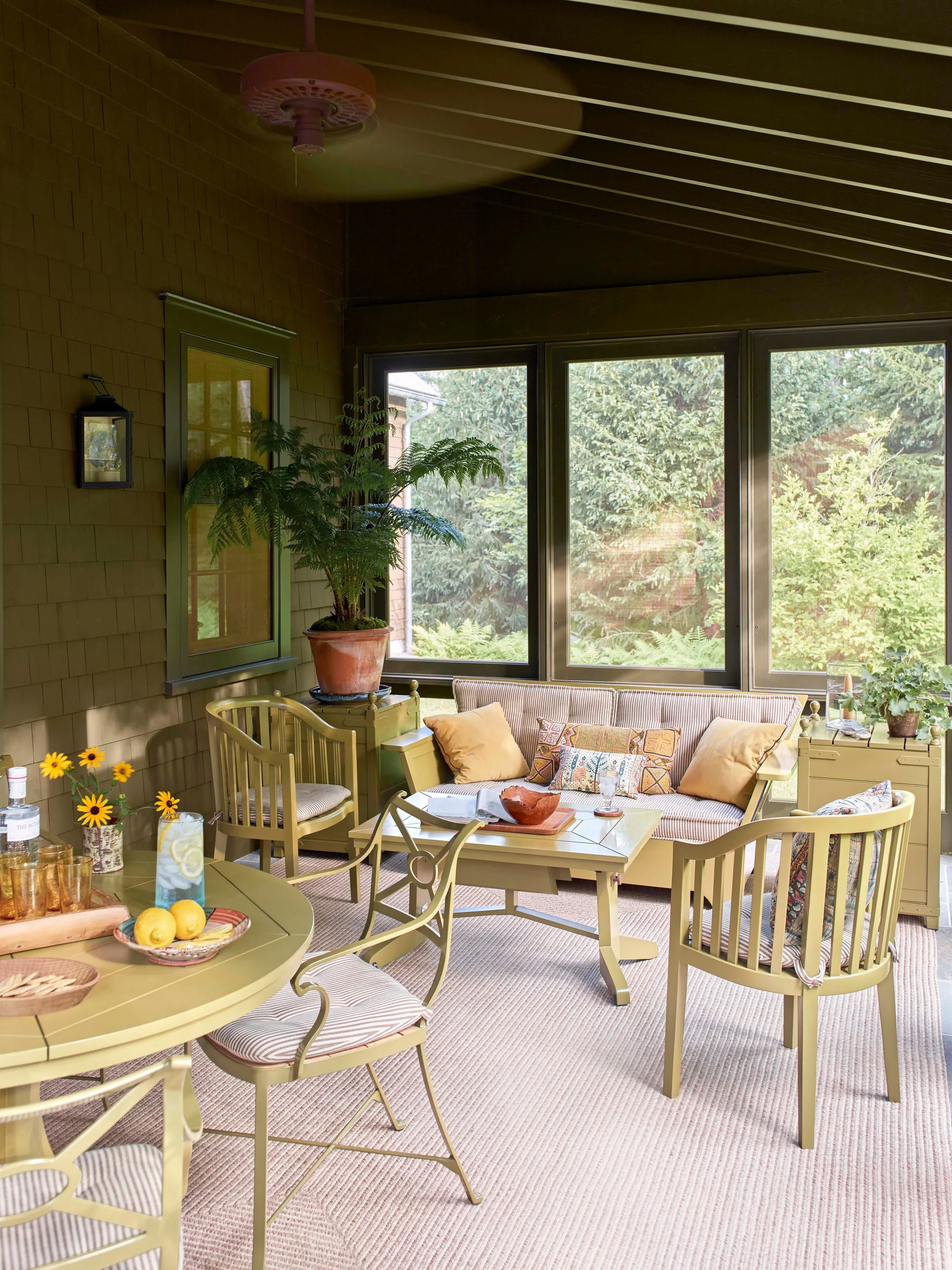 A sunlit screened porch features matching olive green painted wood furniture, striped upholstery, and large windows overlooking trees.