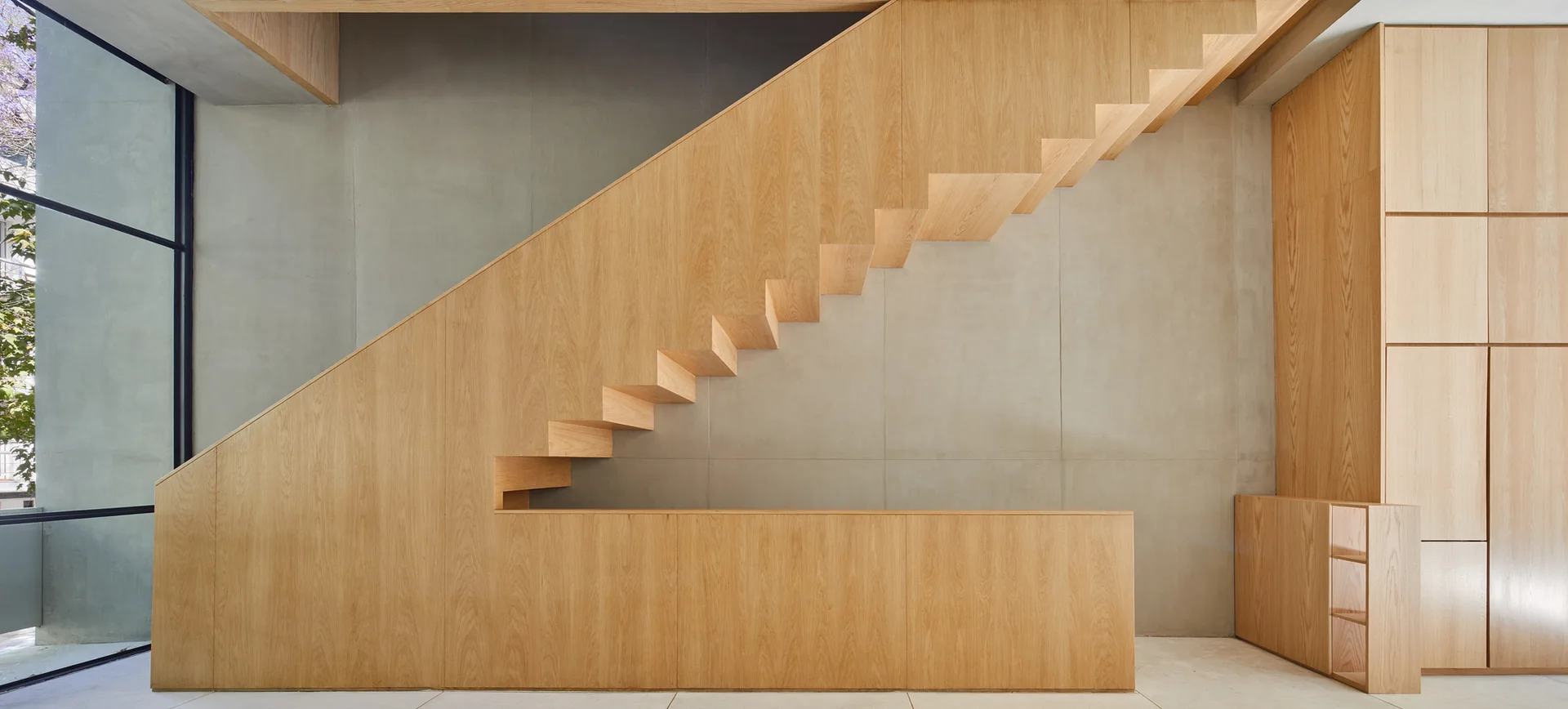 A modern staircase made of light wood with integrated storage cabinets stands against a gray concrete wall next to a black-framed window.