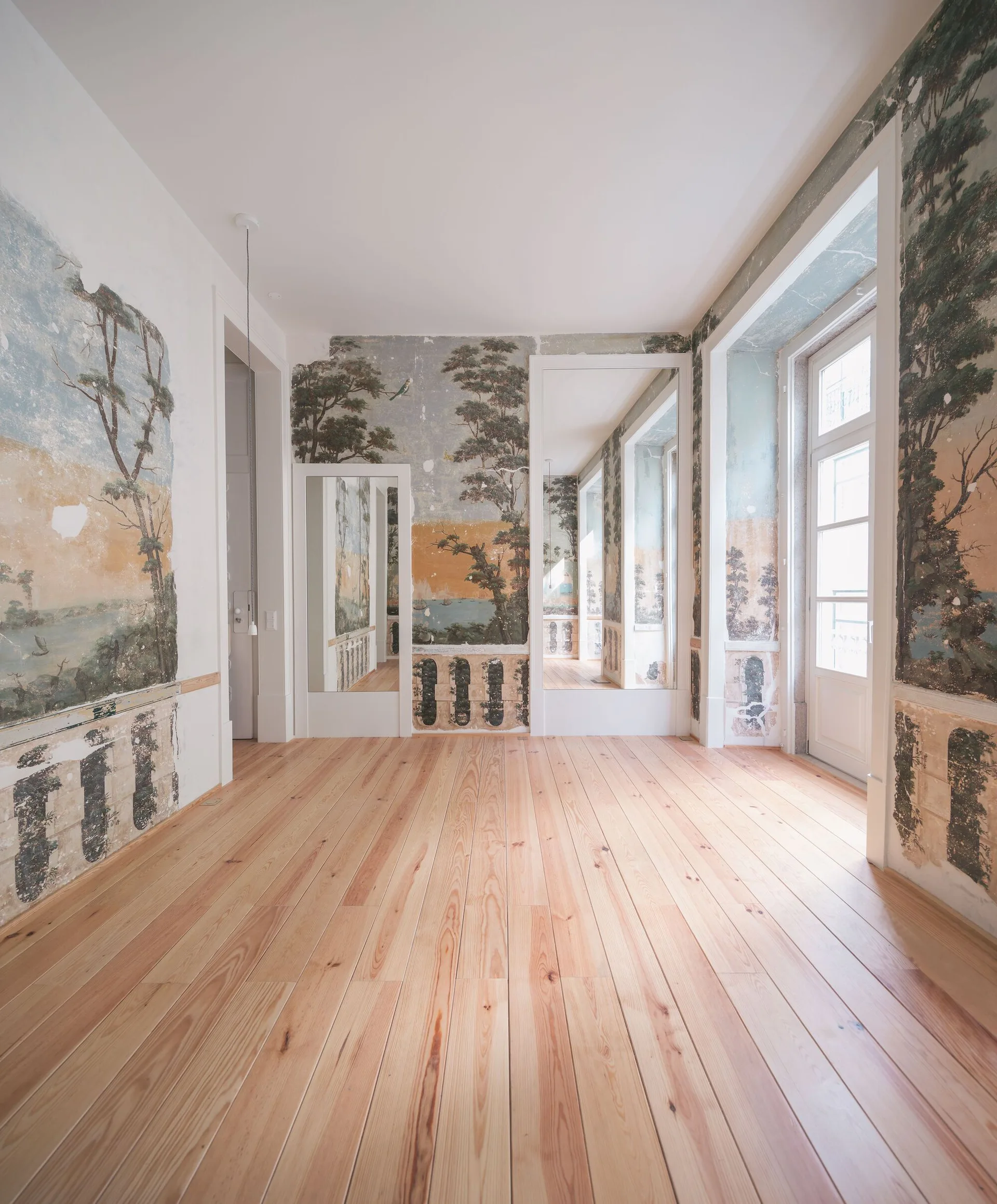 An empty room featuring light wood plank floors and faded landscape murals on the walls with tall windows and inset mirrors.