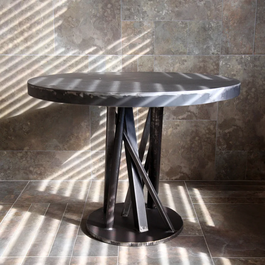 A round dark metal table with an angled, sculptural base stands on a stone tile floor in front of a matching stone tile wall.