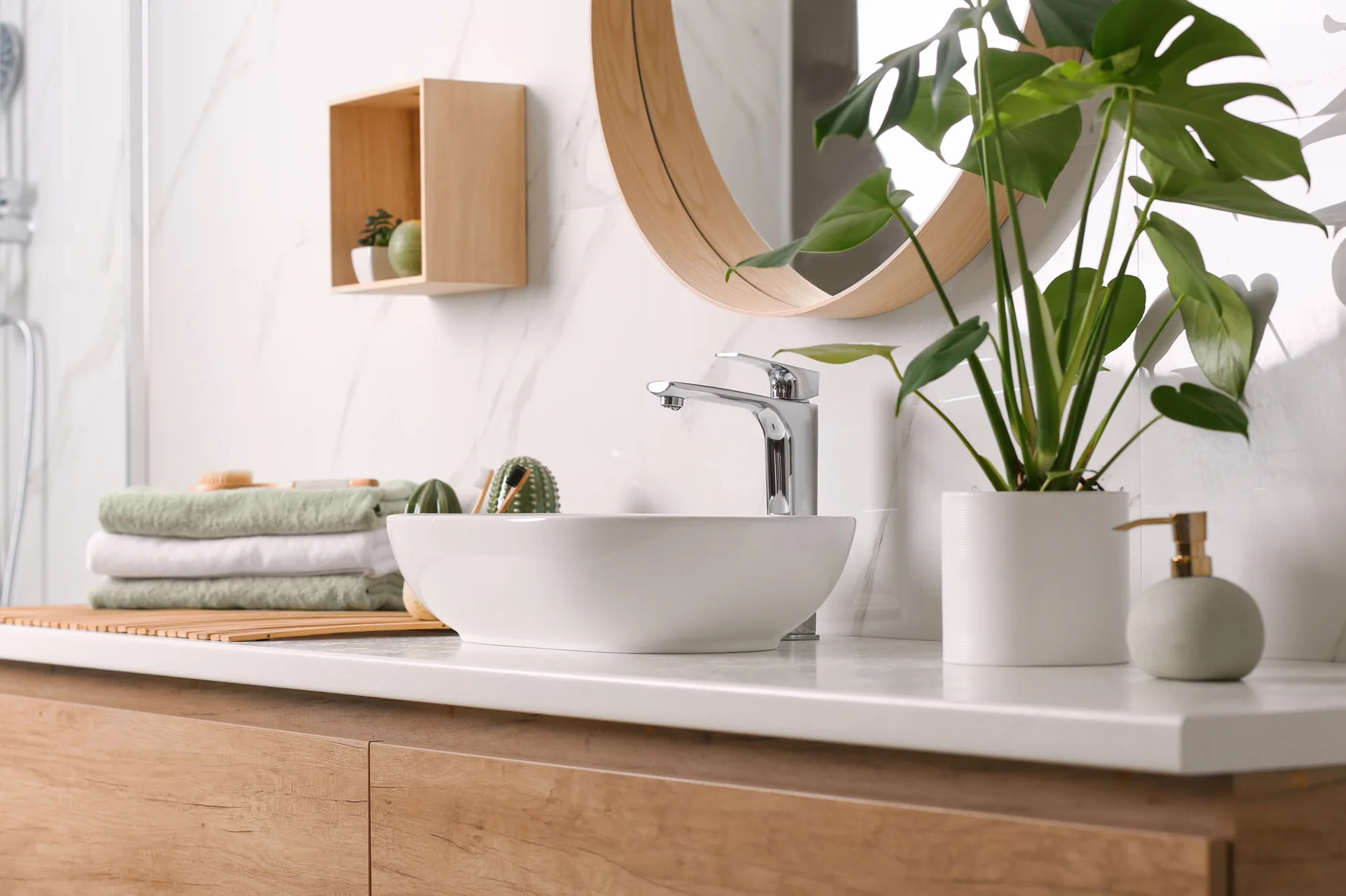 A light wood bathroom vanity featuring a white ceramic vessel sink, a chrome faucet, a round wood-framed mirror, and a small potted plant.