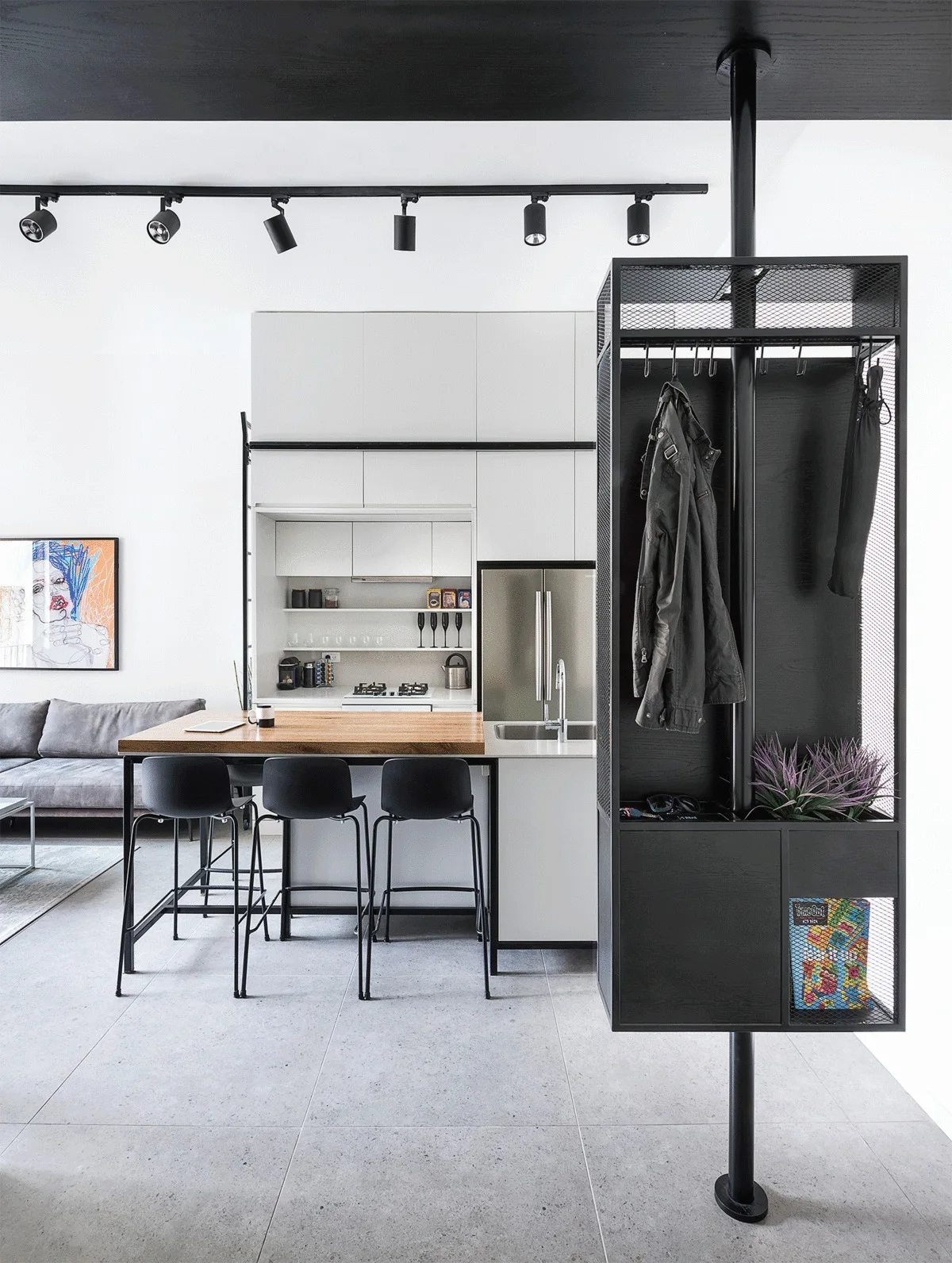 A modern open kitchen features a wood-topped island with black bar stools, white minimalist cabinets, and a tall black metal mesh storage rack.