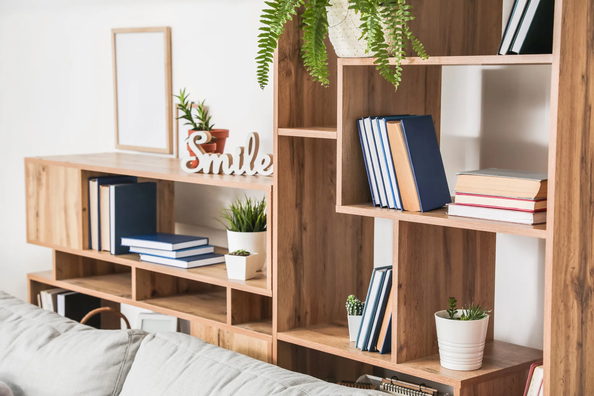 A light wood modular bookshelf styled with books, potted green plants, and decor stands next to a grey fabric sofa against a white wall.