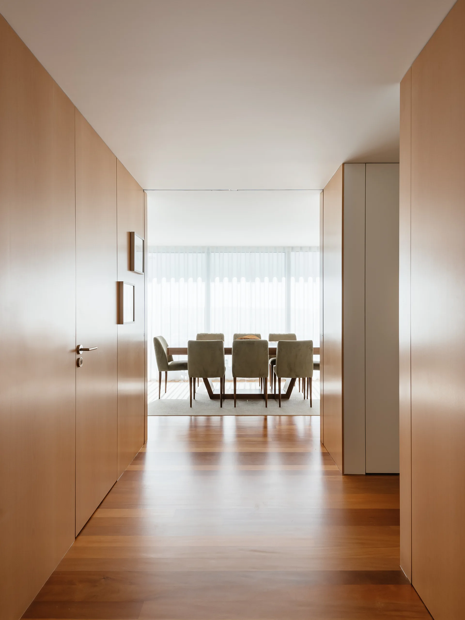 A view through a wood-paneled hallway with wood floors leading to a bright dining room featuring a table, upholstered chairs, and sheer curtains.
