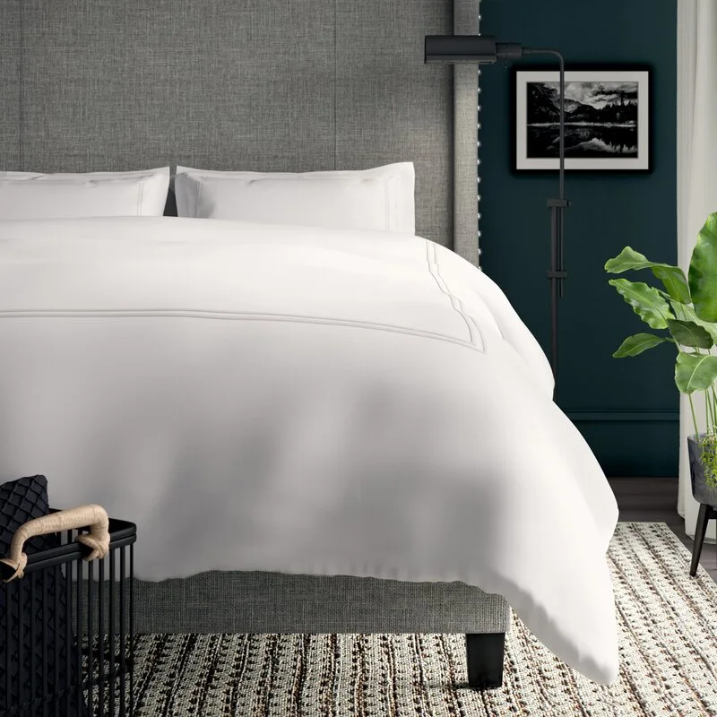 An upholstered grey headboard with white bedding is set against a dark teal wall next to a black floor lamp and a green plant.