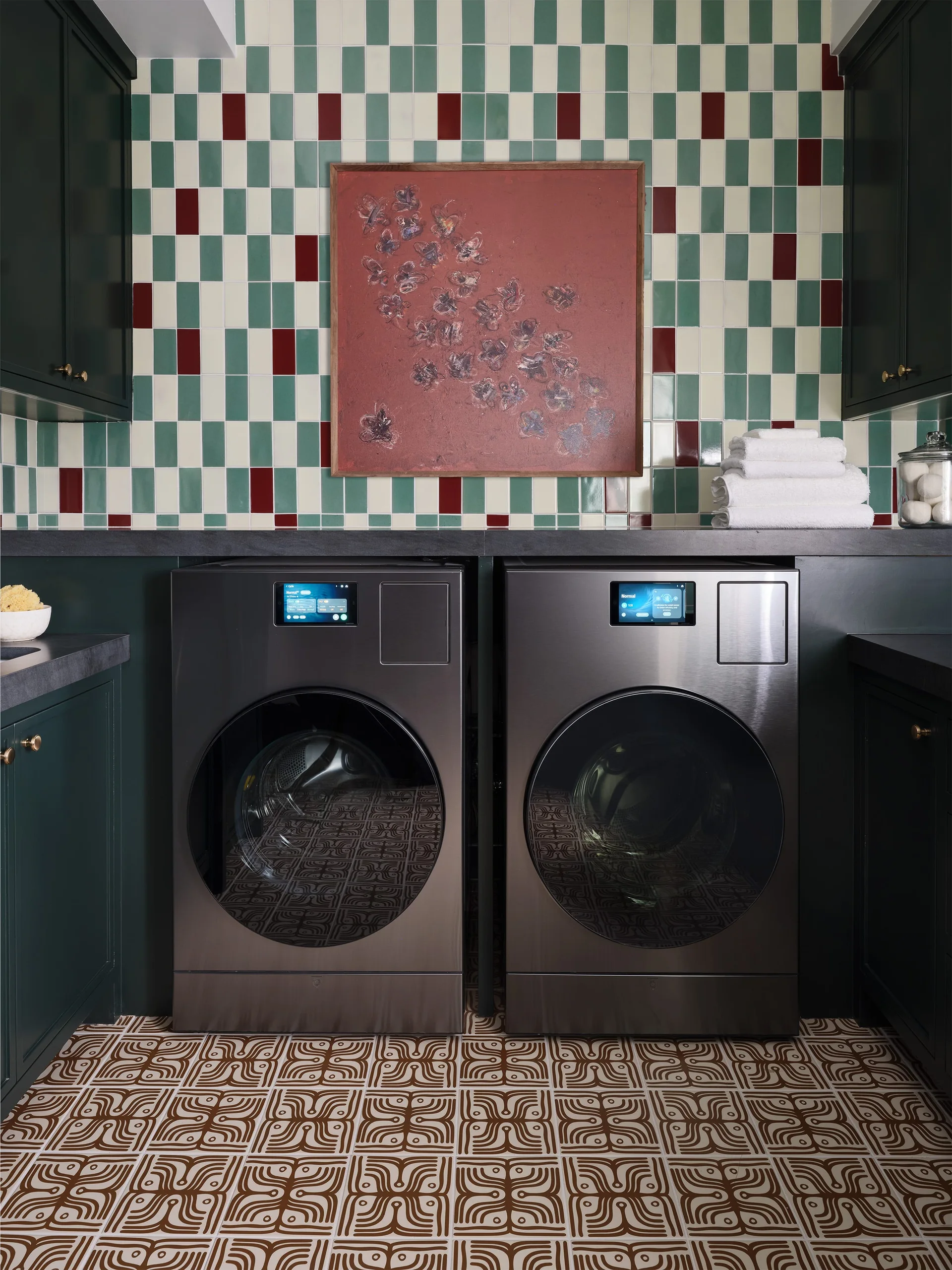 Two dark metallic laundry machines sit beneath a green and white tiled wall with a central reddish framed painting and patterned tile flooring.