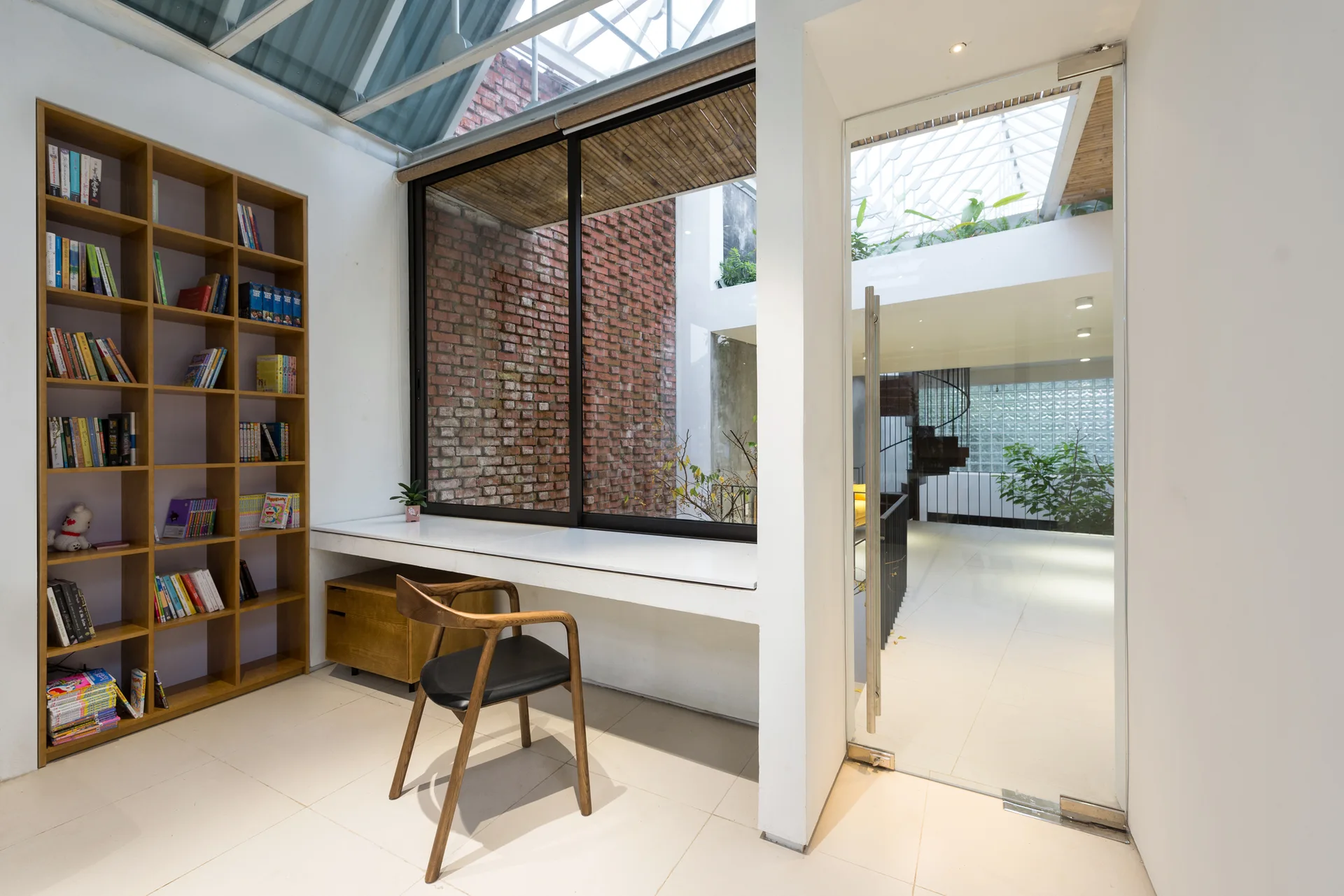A modern home office features a white built-in desk under a glass skylight, alongside a wooden bookshelf and a transparent glass door partition.