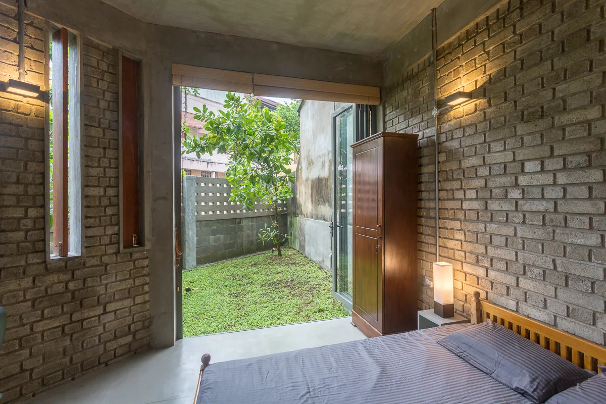 An industrial bedroom features exposed brick walls, a wooden bed, and a large open glass door leading to a grassy courtyard.