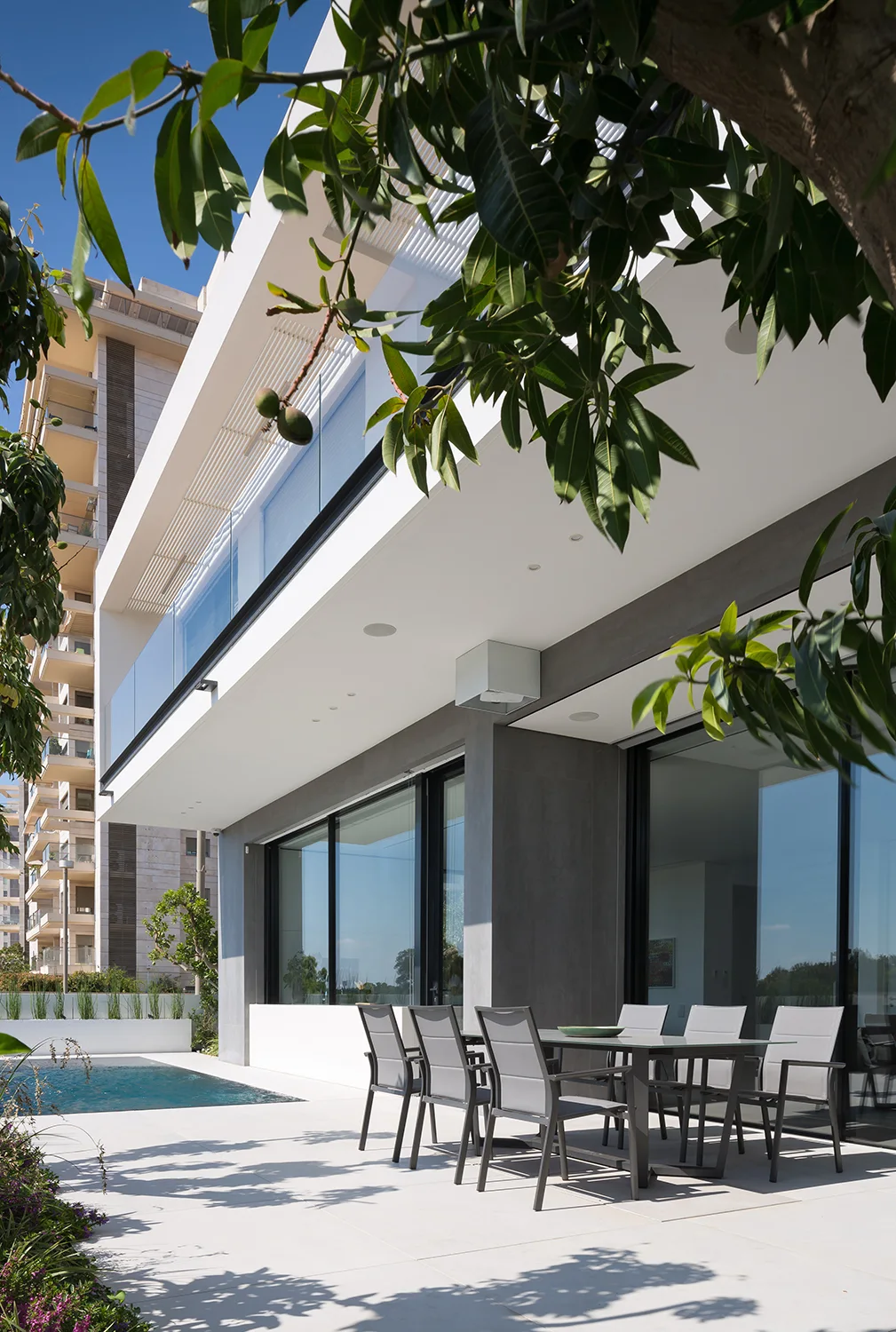 A modern grey dining set sits on a white tiled patio next to a swimming pool under a deep architectural overhang.