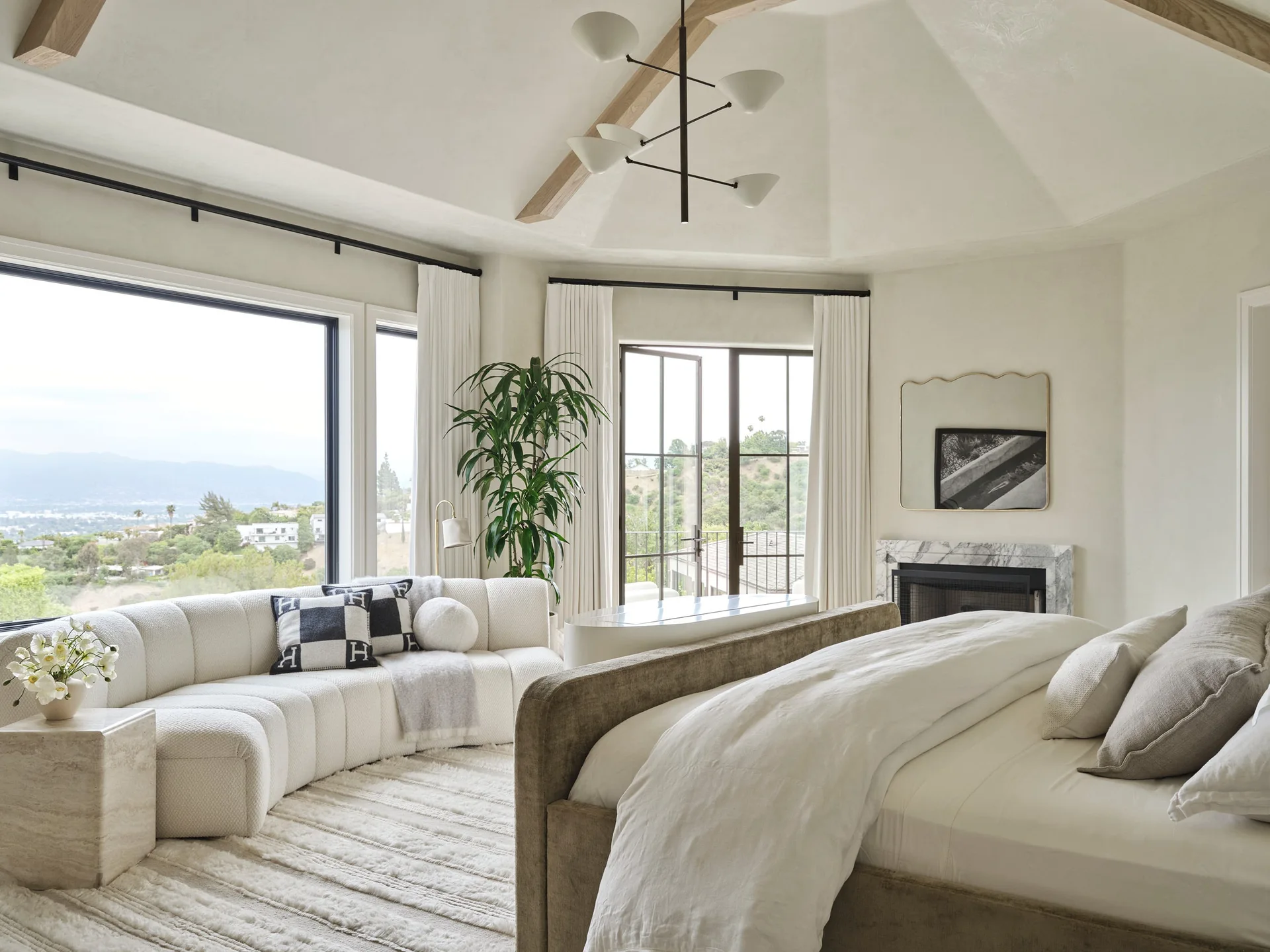 A bright bedroom features an upholstered bed in the foreground, a curved cream sofa by large windows, a marble fireplace, and a vaulted ceiling.