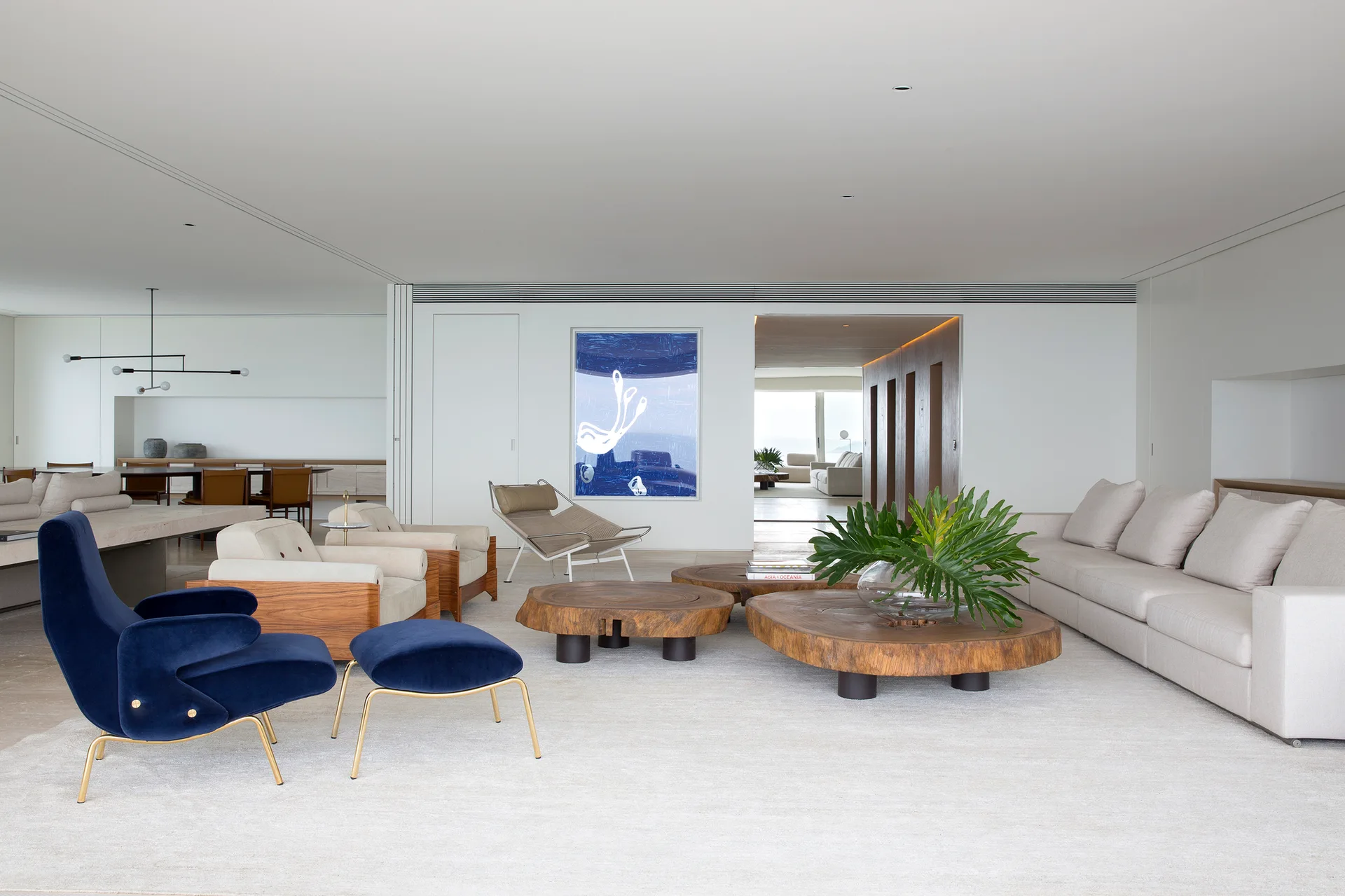 A spacious minimalist living room with a large cream sofa, organic wood slab coffee tables, and a vibrant blue velvet armchair on a white rug.