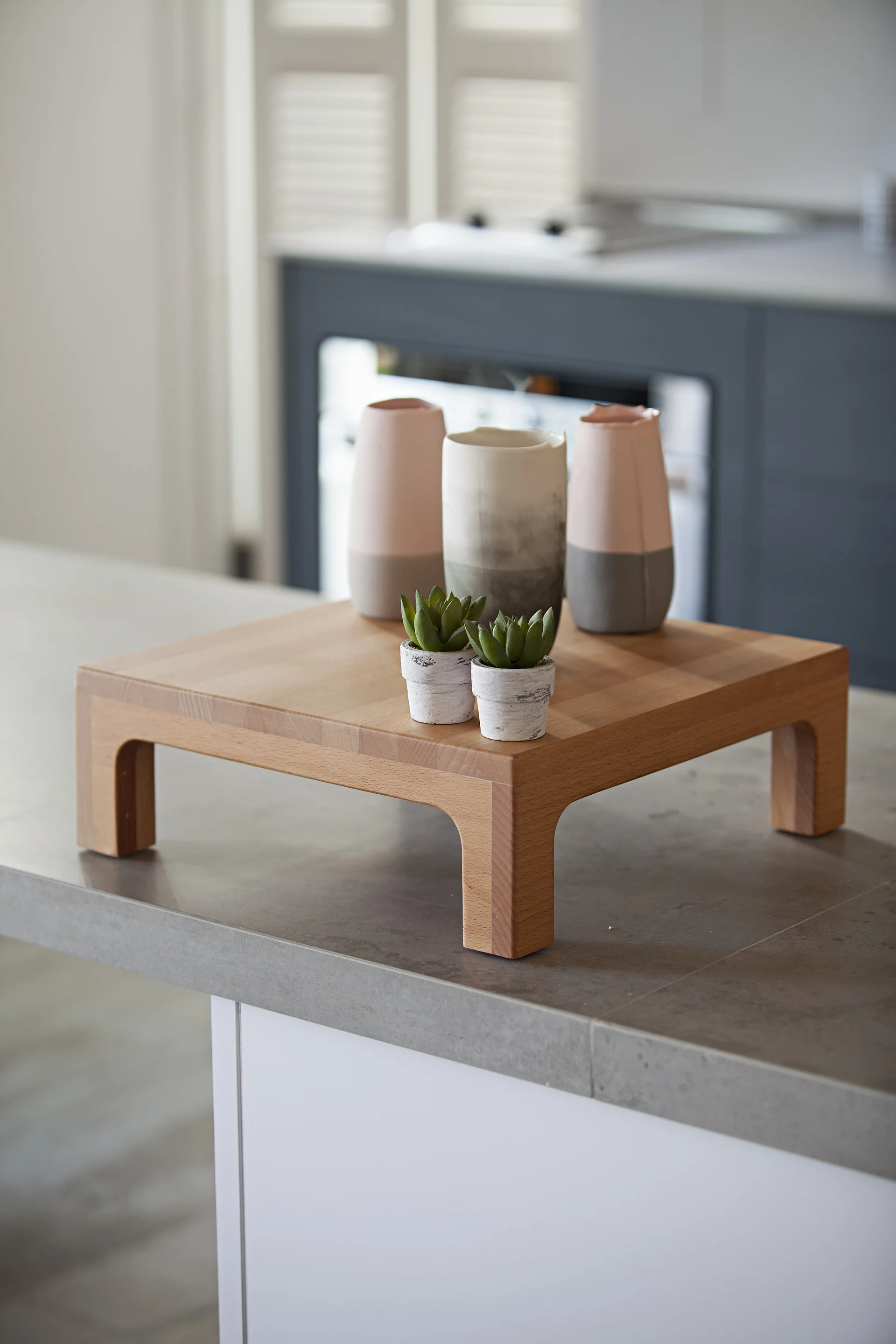 A low wooden pedestal on a gray countertop holds three ceramic vases and two small succulents.