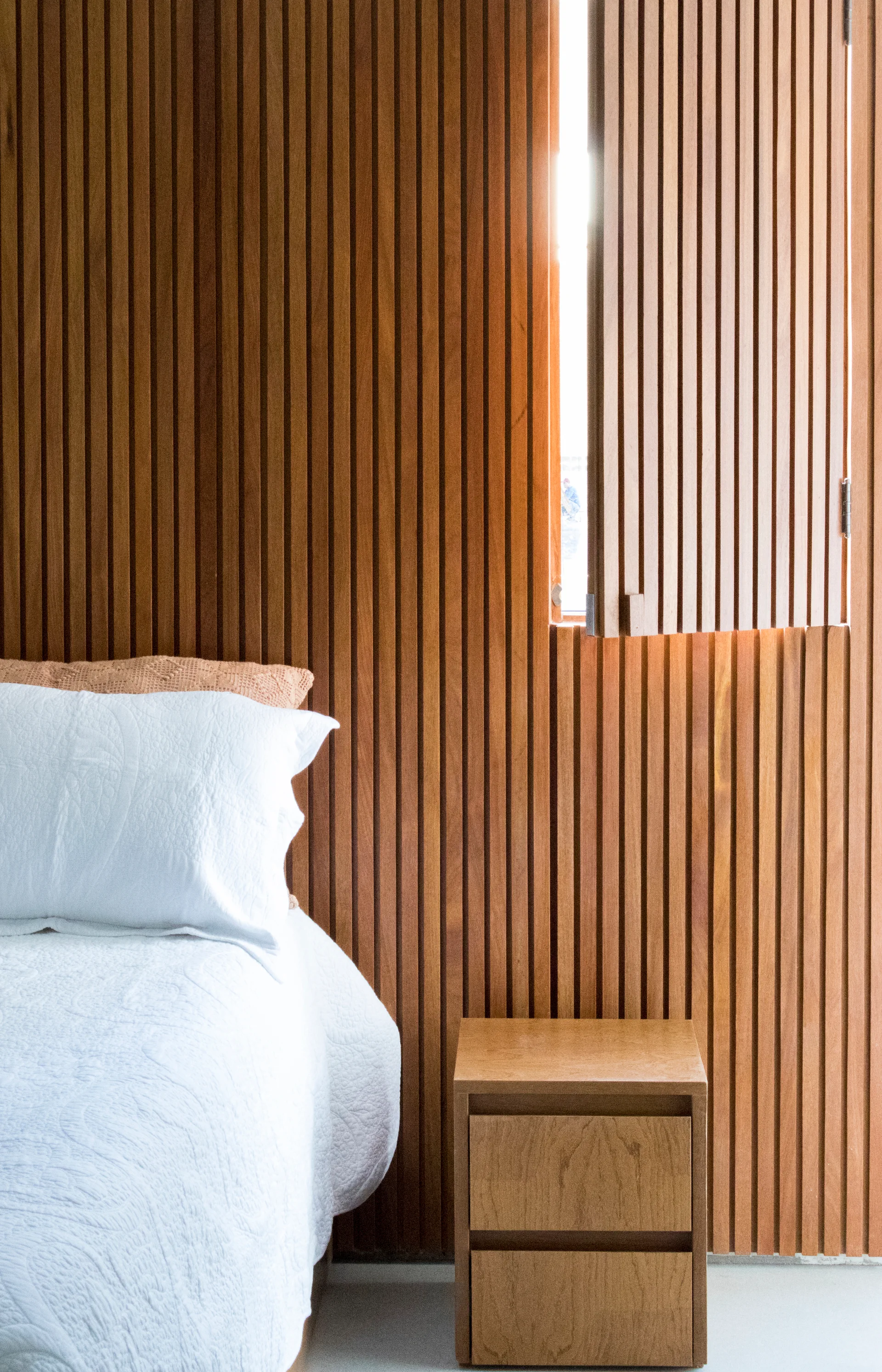 A bedroom corner features a bed with white textured bedding, a wooden nightstand, and a vertical wood slat wall with an integrated open window shutter.