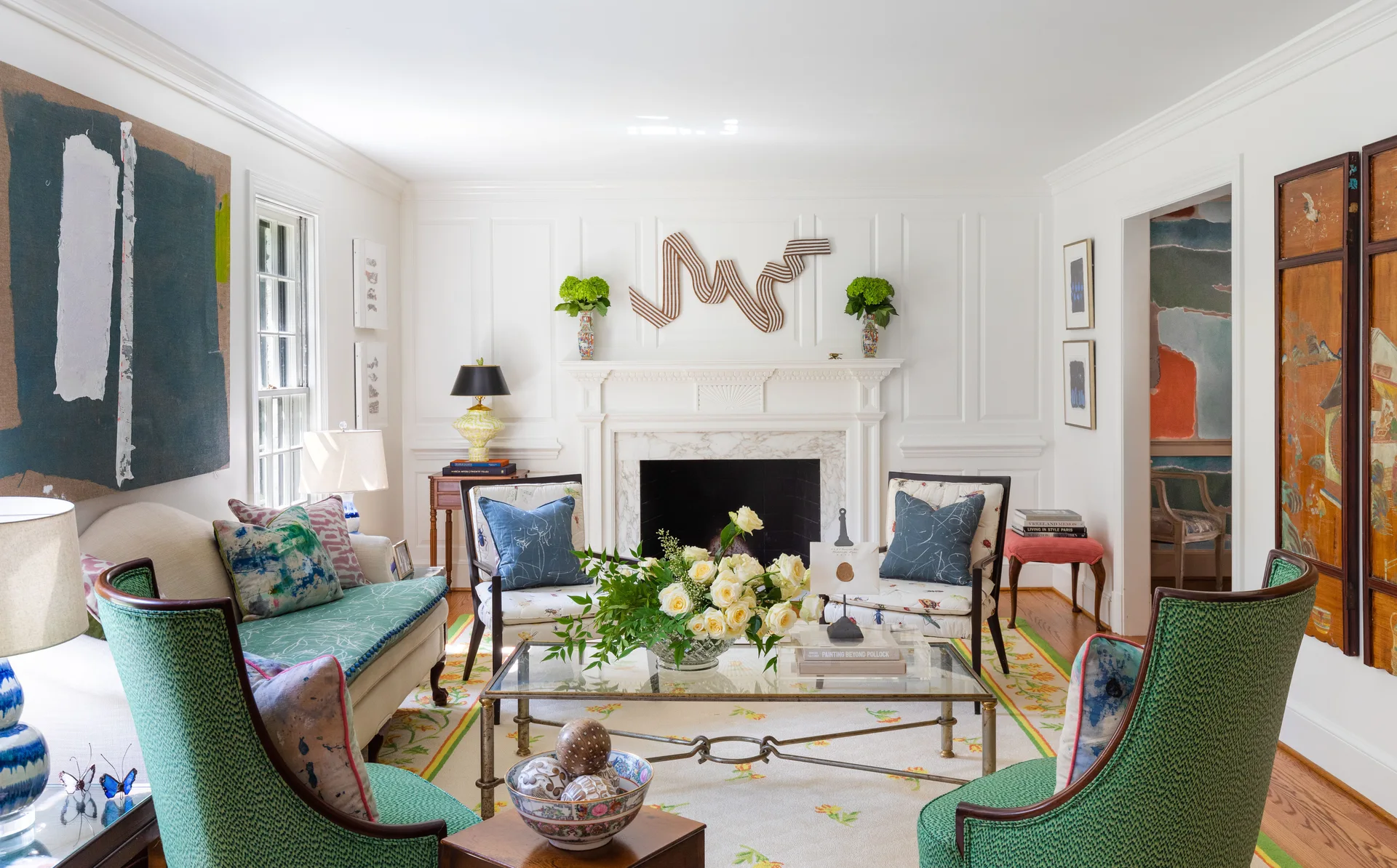 A bright living room features green patterned armchairs, a glass coffee table, and a white fireplace surrounded by wainscoted walls.