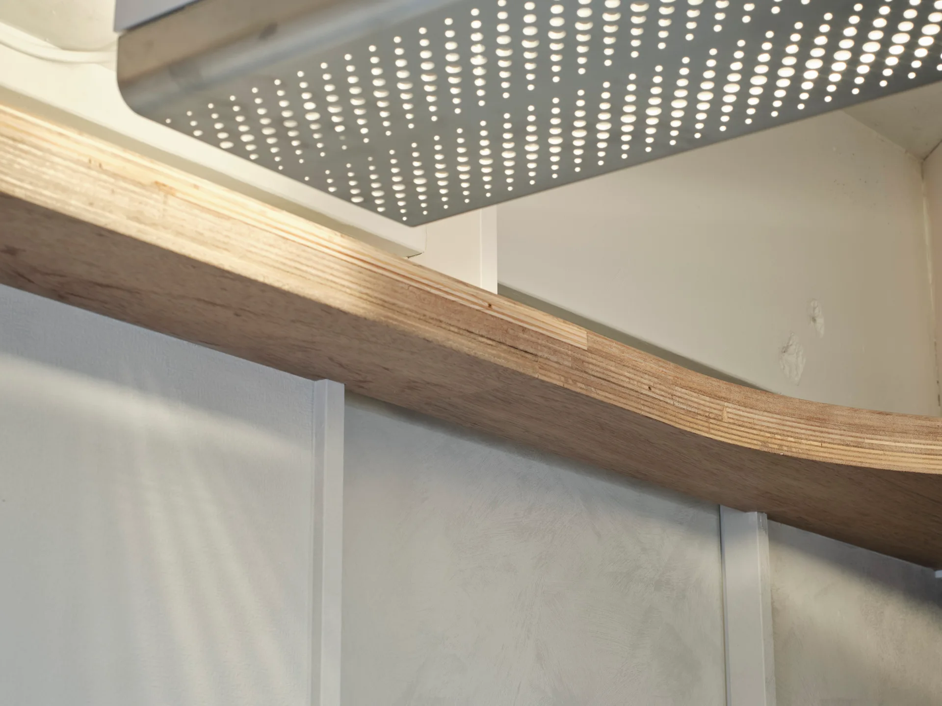 A close-up of a curved plywood shelf beneath a perforated metal light fixture mounted on a white wall.