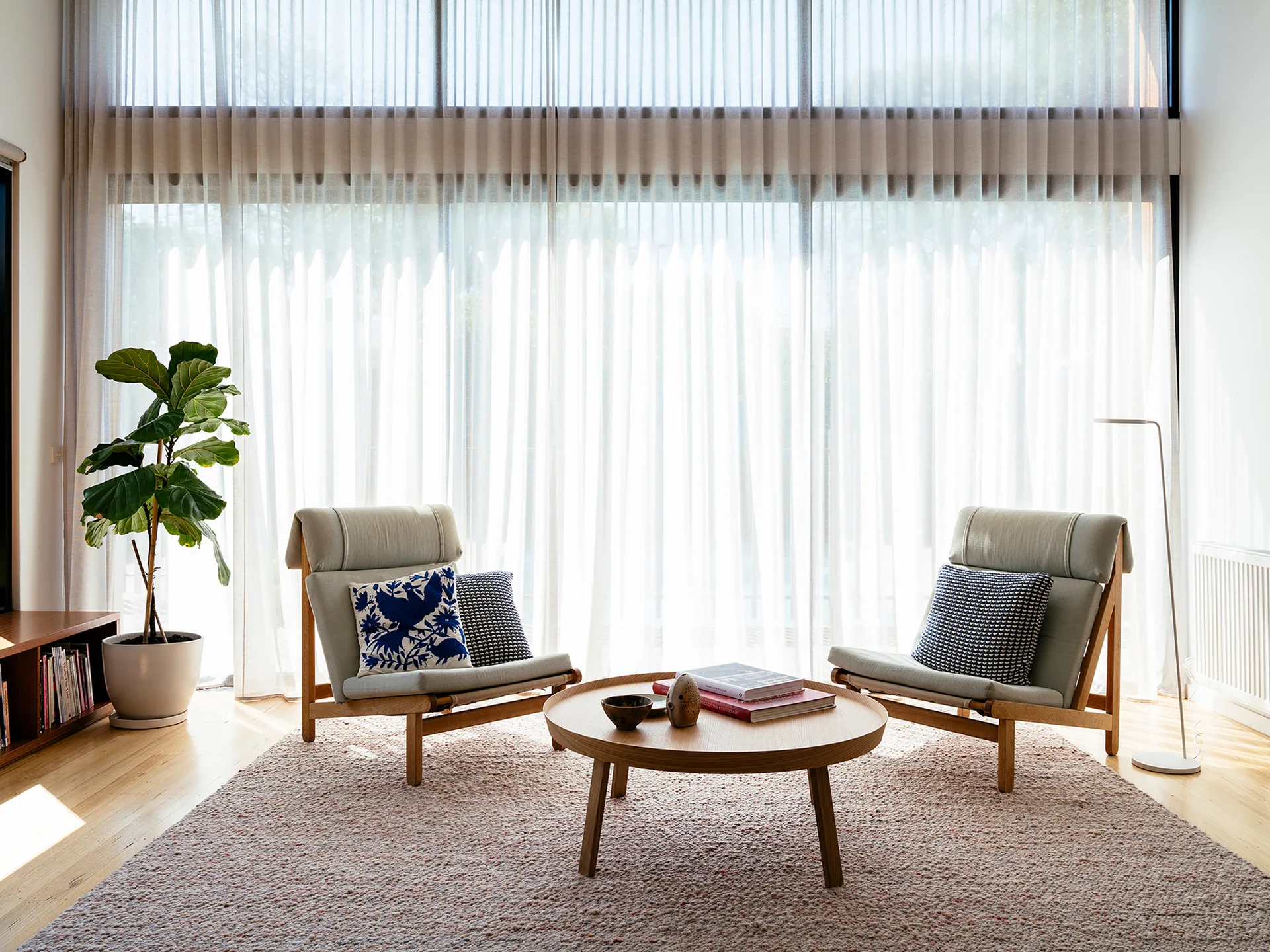Two grey Scandinavian armchairs stand on a textured beige rug around a round wooden coffee table in front of floor-to-ceiling sheer curtains.
