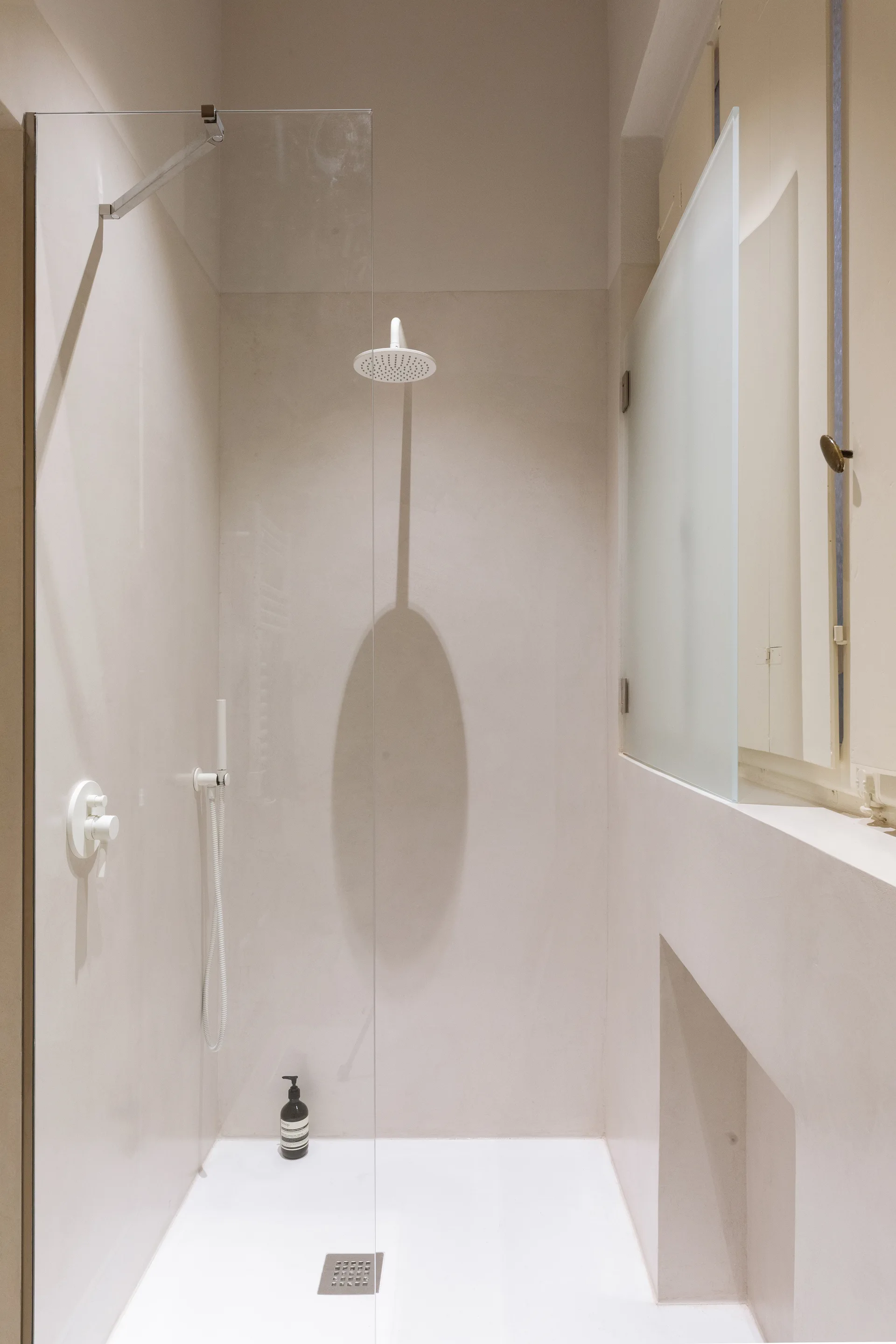 A minimalist shower stall features seamless beige plaster walls, white overhead and handheld fixtures, and a clear glass panel partition.