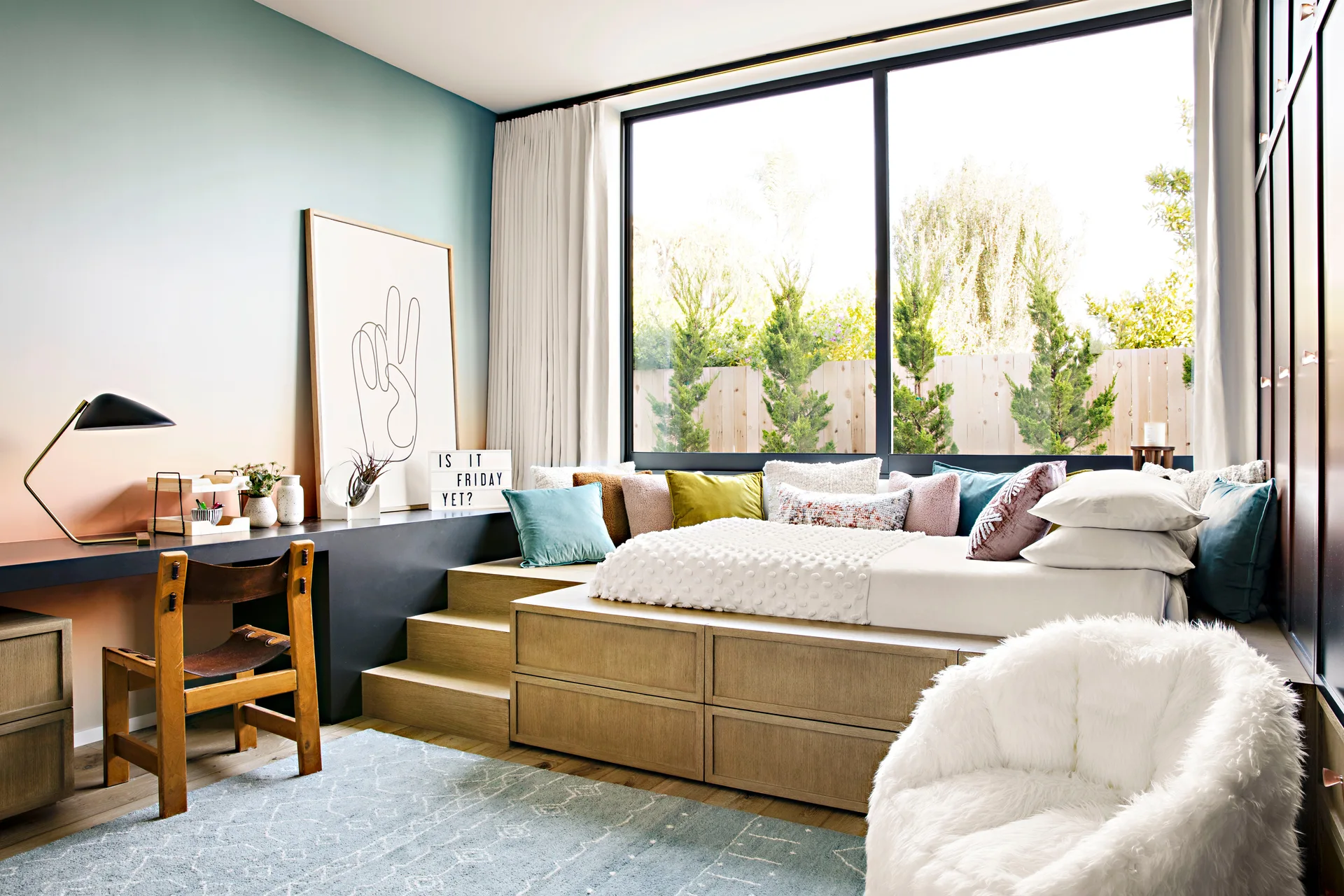A modern kids bedroom features an elevated wooden platform bed with stairs, an integrated desk, and a colorful peach-to-teal ombre wall.