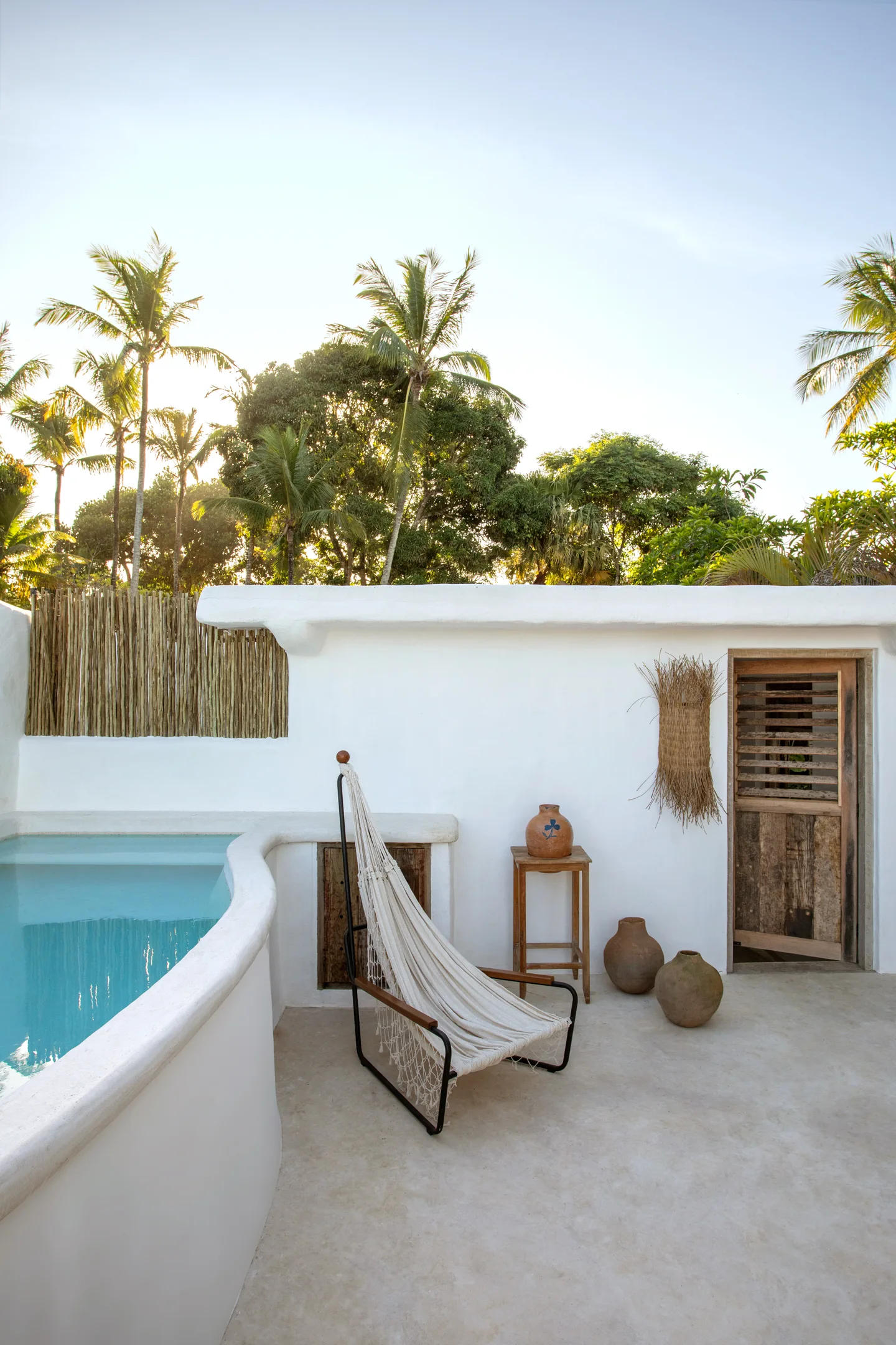 A white hammock chair sits on a concrete patio next to a curved swimming pool and a rustic plastered wall decorated with ceramic jars.