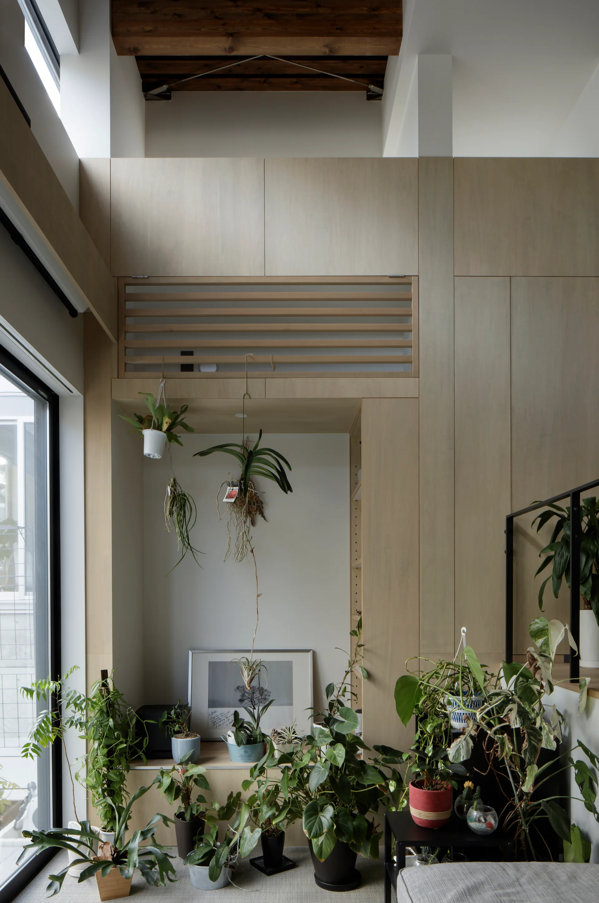 A light wood built-in cabinet wall features a recessed niche filled with numerous potted houseplants, hanging orchids, and a framed art print.