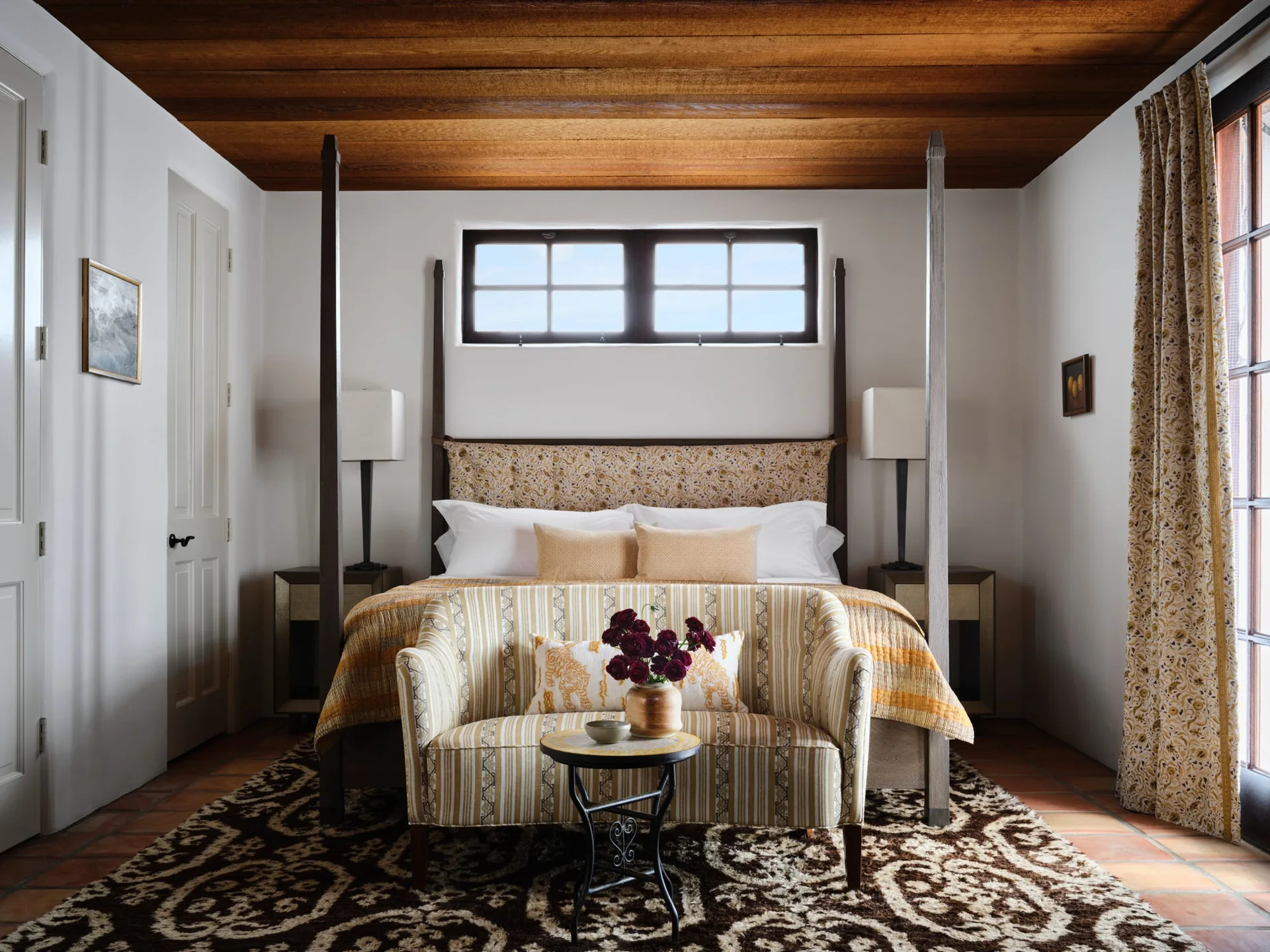A bedroom features a dark wood poster bed beneath a wood plank ceiling with a striped loveseat placed at the foot on a patterned rug.