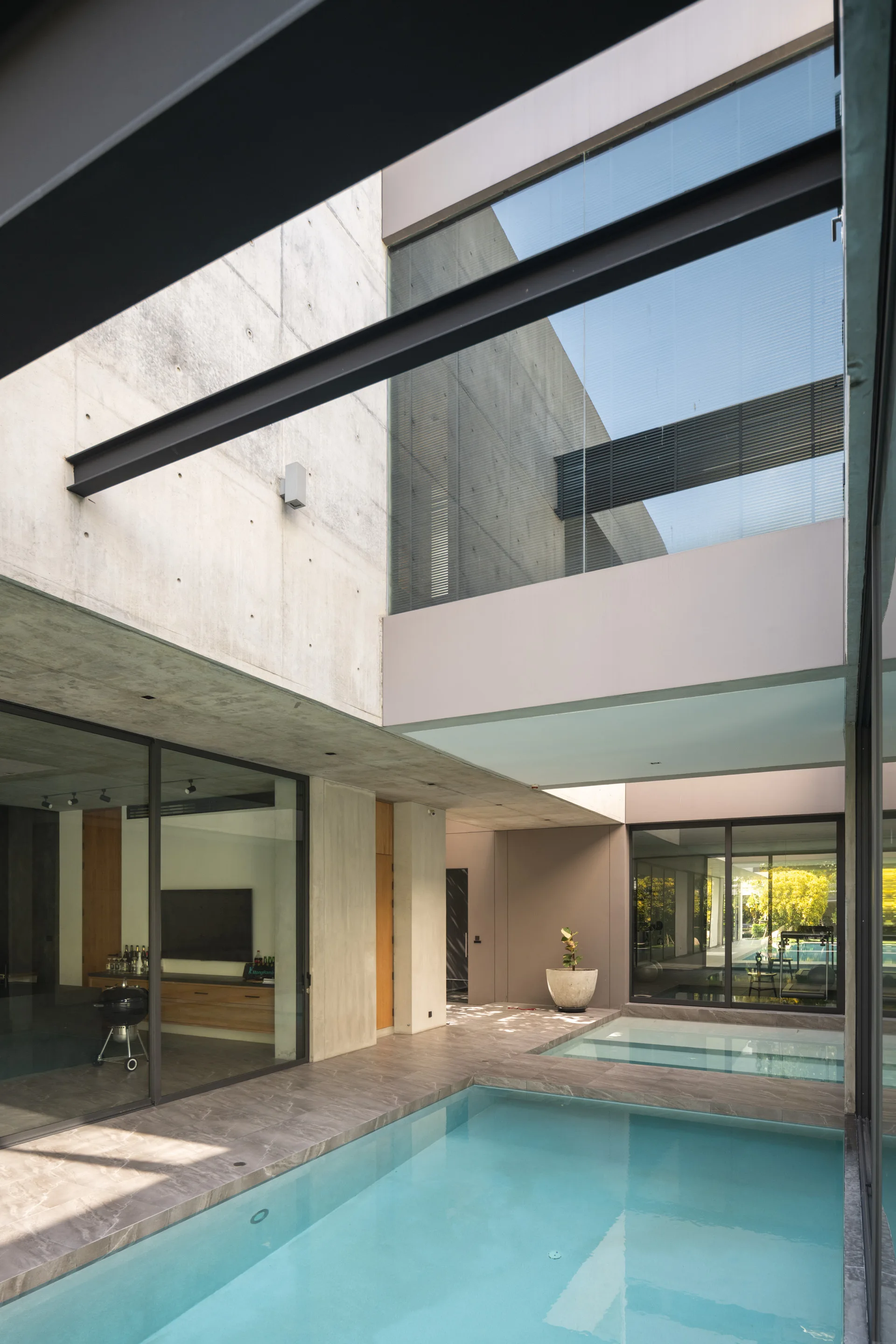 A minimalist outdoor courtyard features a turquoise swimming pool surrounded by grey stone tiles, exposed concrete walls, and large glass windows.