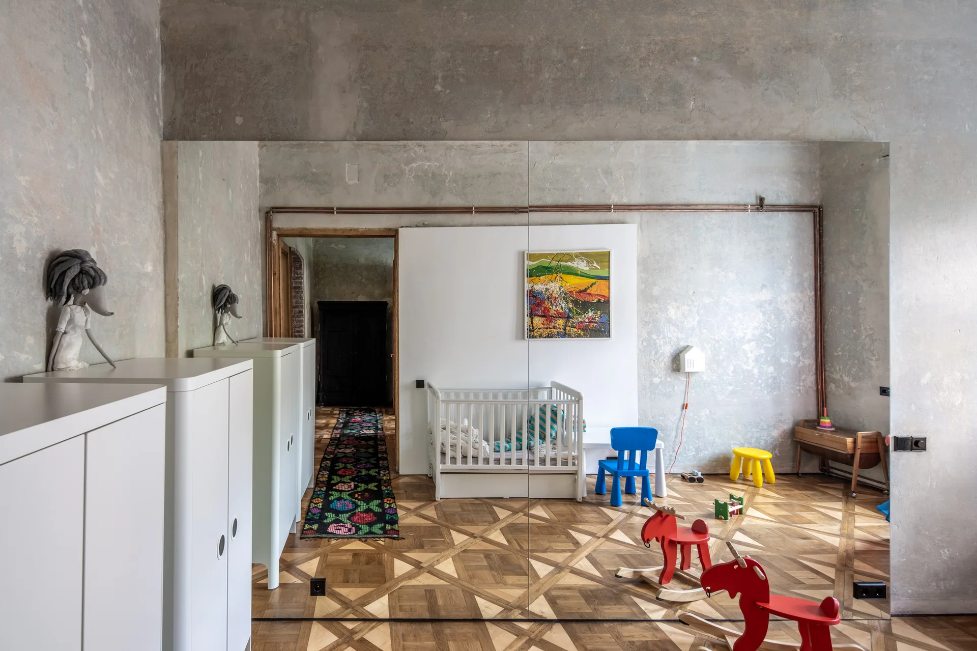 A modern children's nursery featuring geometric wood parquet flooring, distressed plaster walls, a white crib, and a mirrored wall.