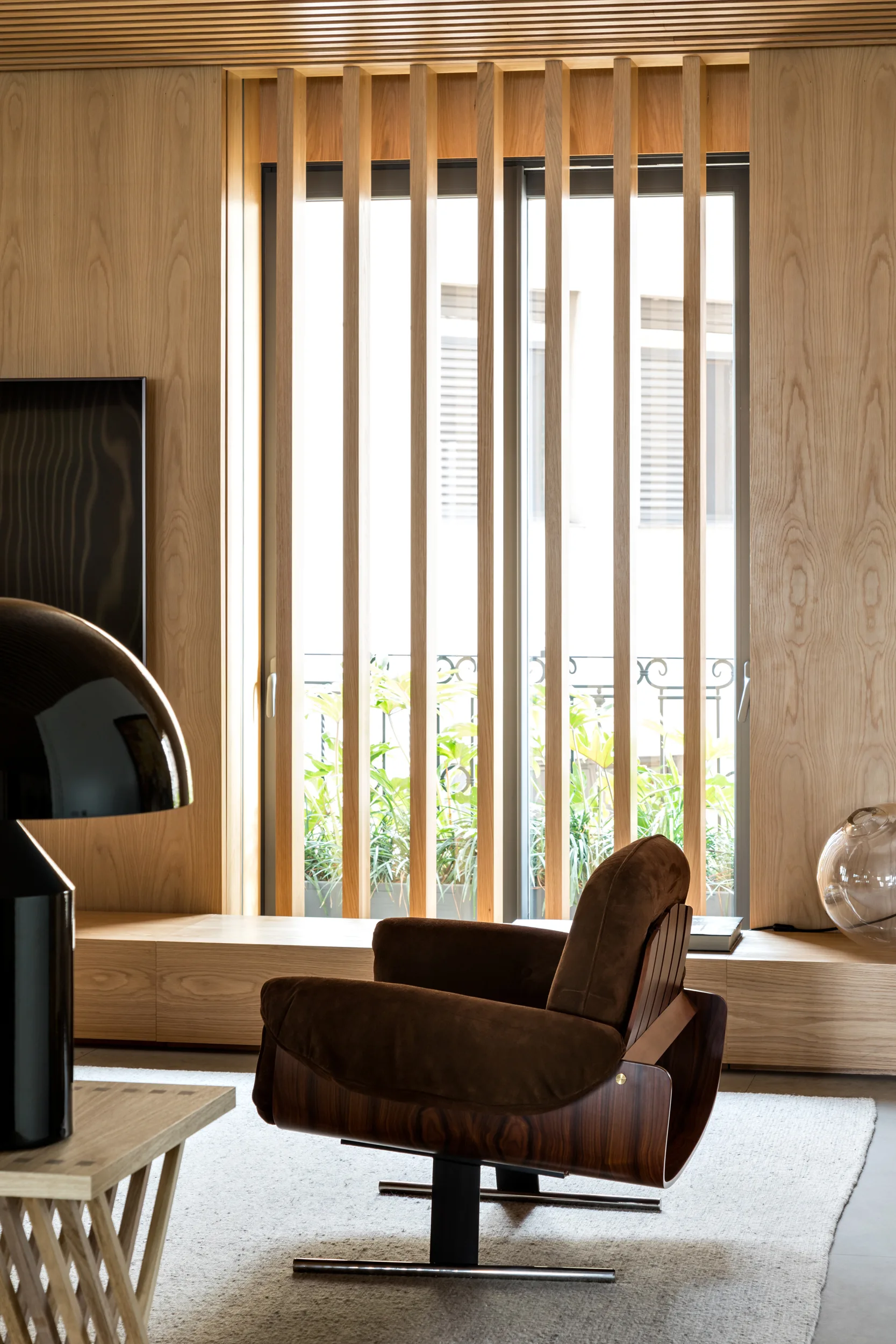 A modern living room features light wood wall paneling, a large window filtered by vertical wood slats, and a brown upholstered armchair on a beige rug.