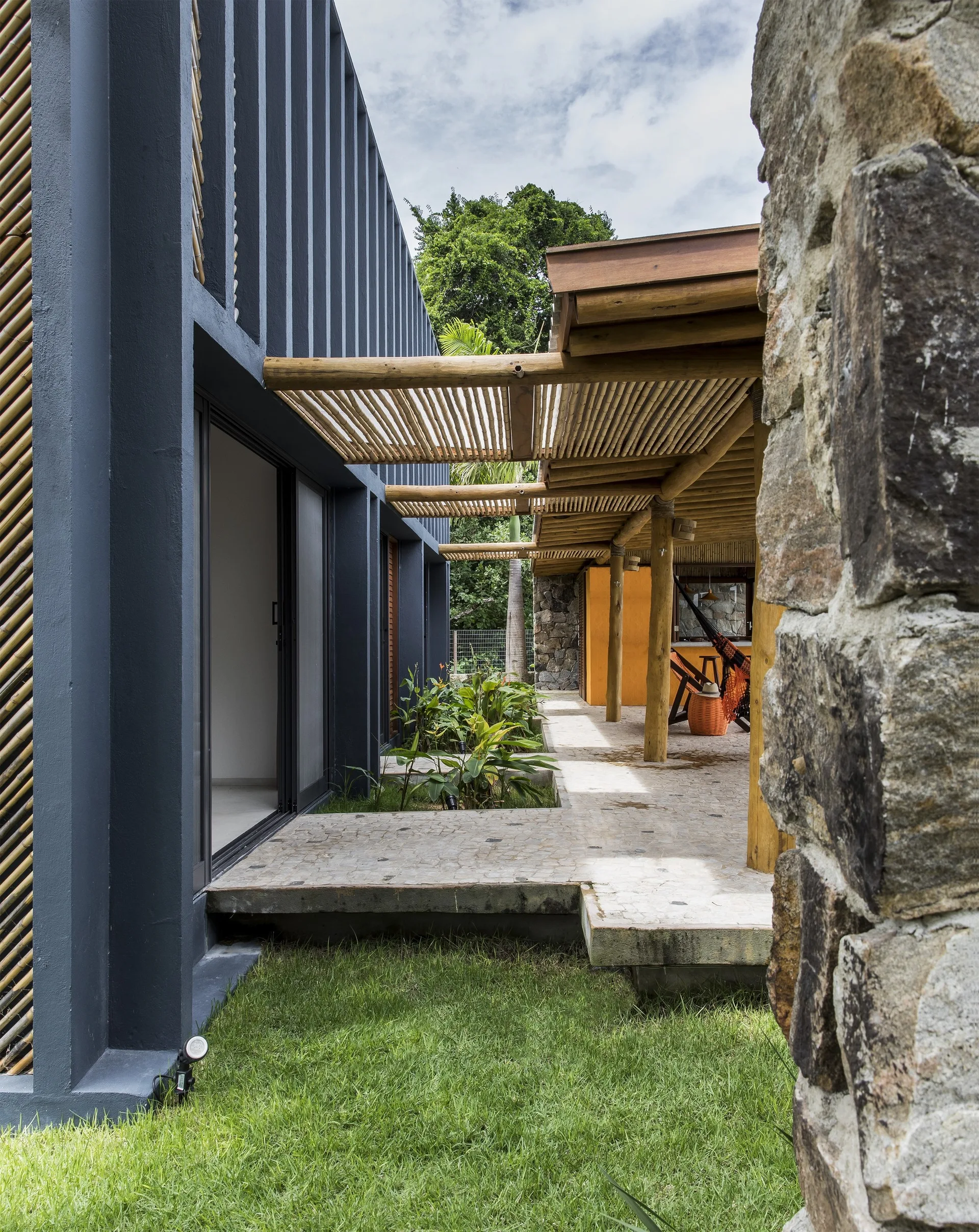 A modern outdoor patio features a bamboo slatted pergola attached to a dark grey concrete wall with stone columns and tropical plants.