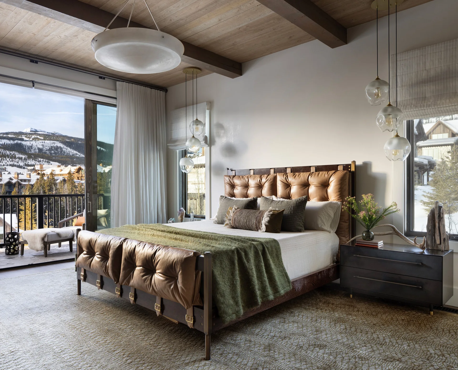 A bedroom features a tufted brown leather bed, wood ceiling beams, hanging glass globe pendants, and a sliding glass door showing a snowy landscape.
